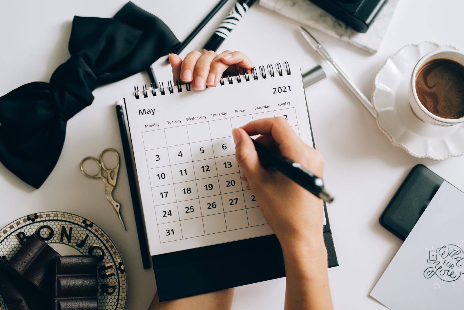 Hands marking dates on a monthly desk calendar with a pen, a cup of coffee nearby.