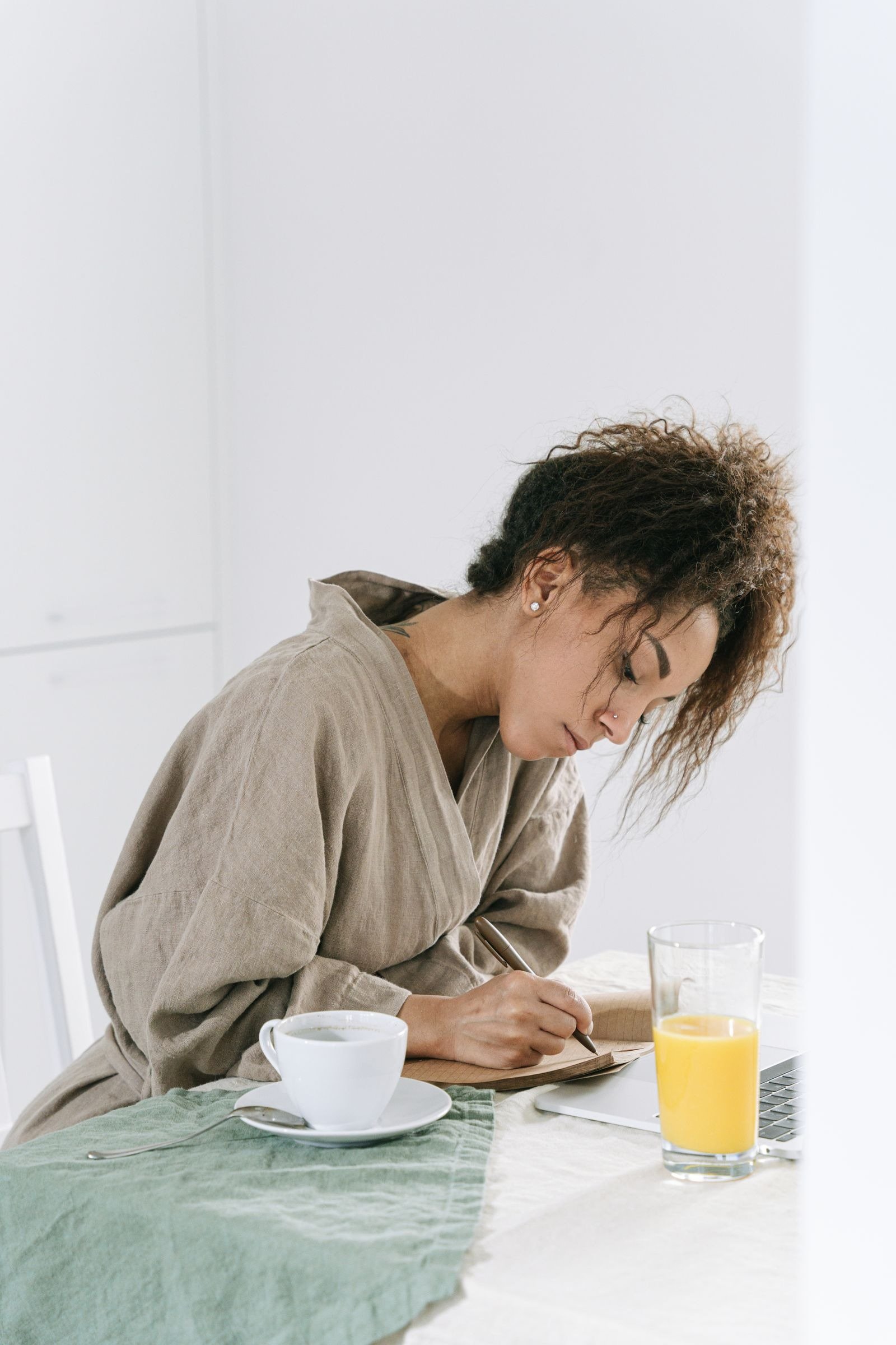 A woman in a robe writing in a notebook at a table with coffee and orange juice.