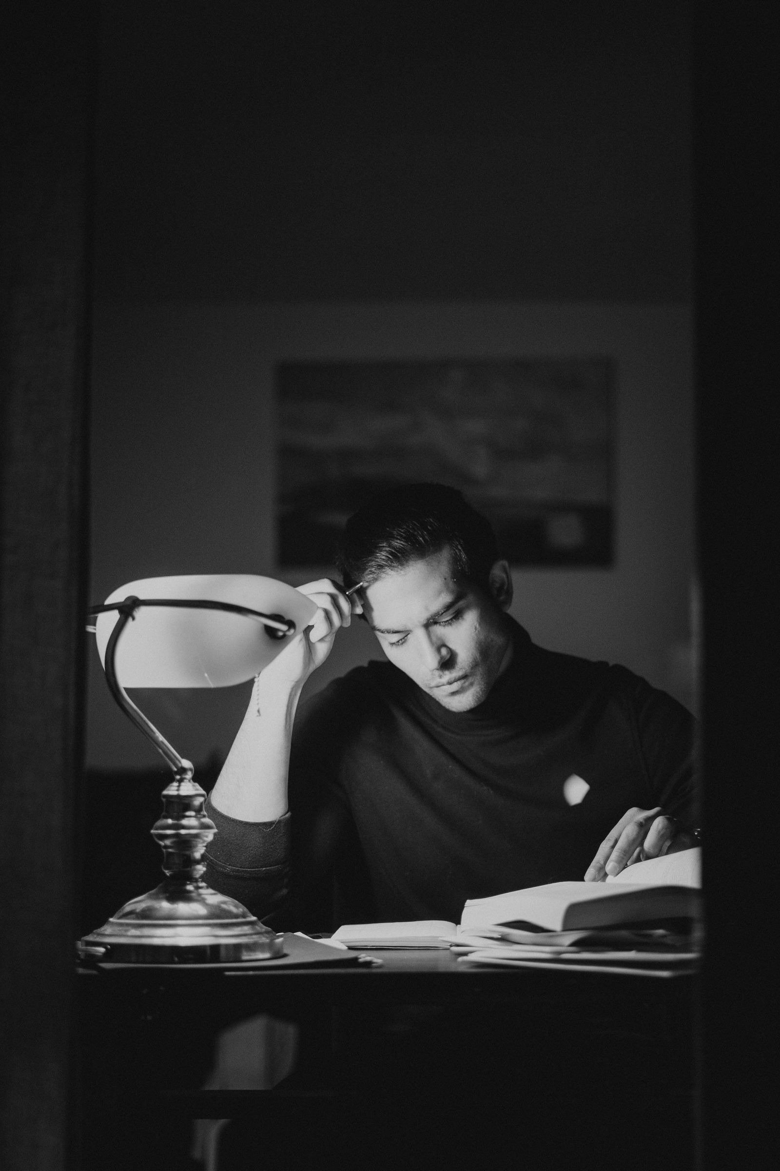 A black-and-white photo of a man studying books at a desk under a lamp late at night.