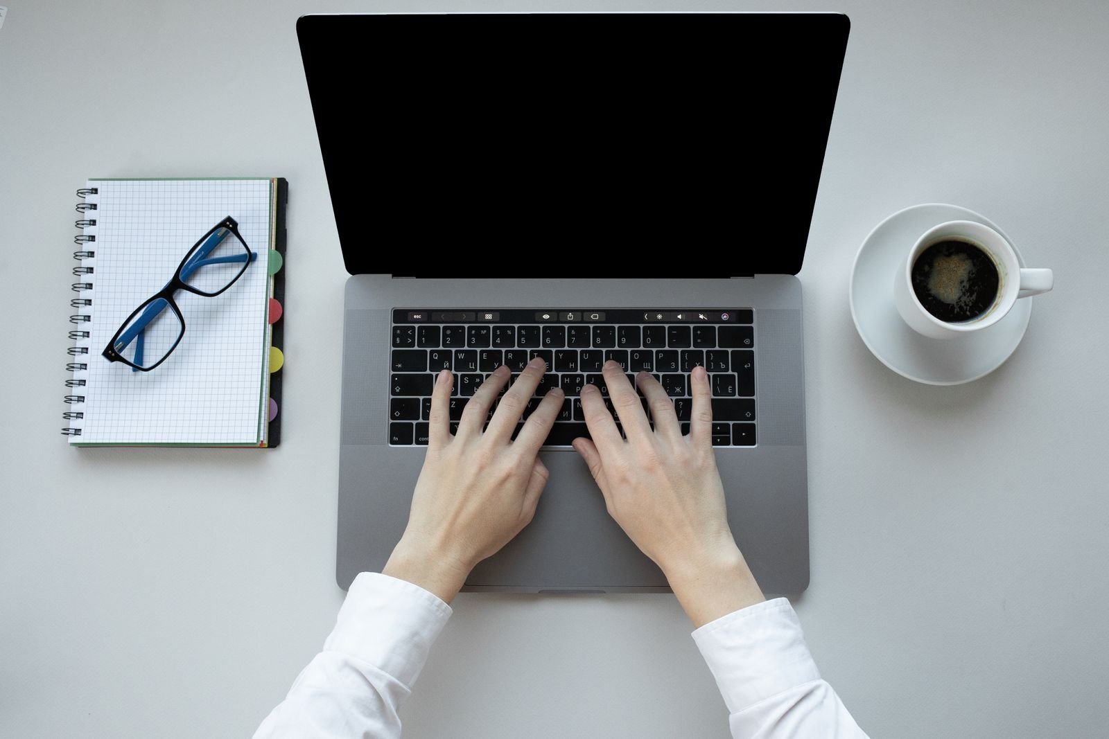 Hands typing on a MacBook laptop with a coffee cup, glasses, and notebook on a white desk.