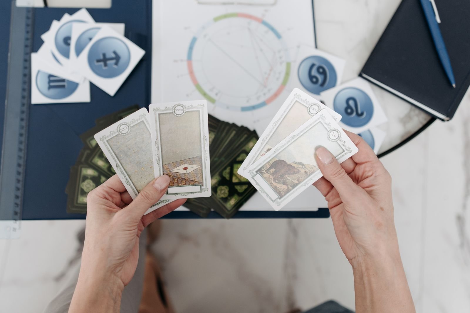 Two hands holding tarot cards over a desk with a printed chart wheel, zodiac symbol cards, a ruler, and a notebook.