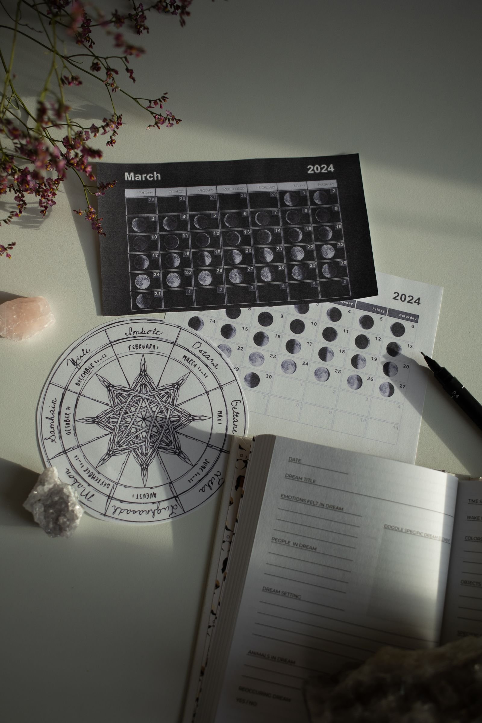 A flat lay with moon-phase calendars, a hand-drawn wheel-of-the-year diagram, an open dream journal, crystals, and dried flowers.