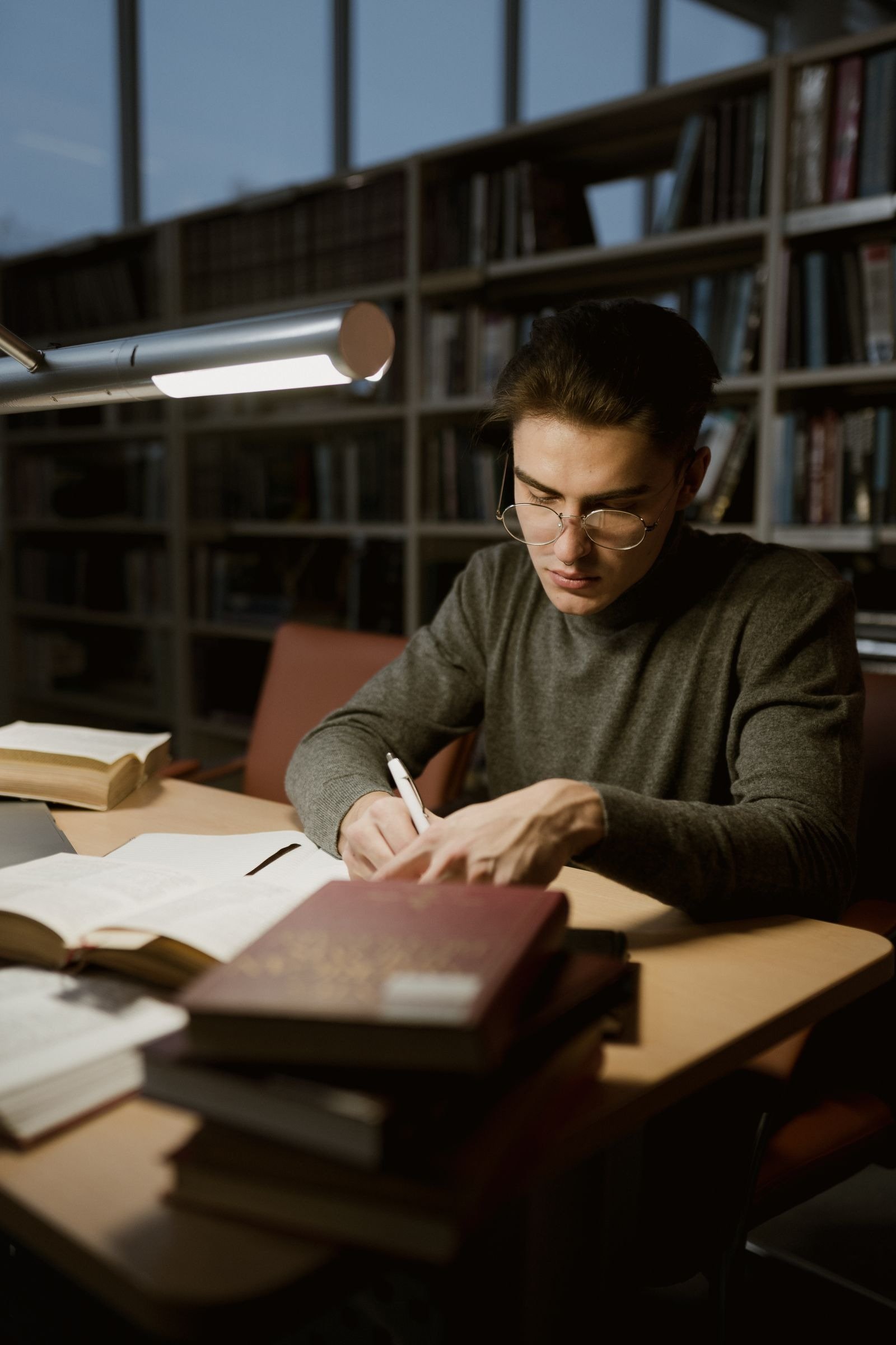 A young man with glasses studying books at a library desk under a lamp.