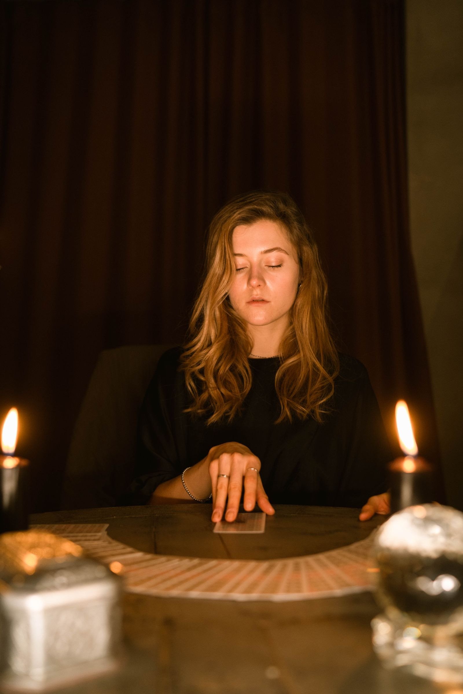 A woman with closed eyes touching a tarot card on a candlelit table.