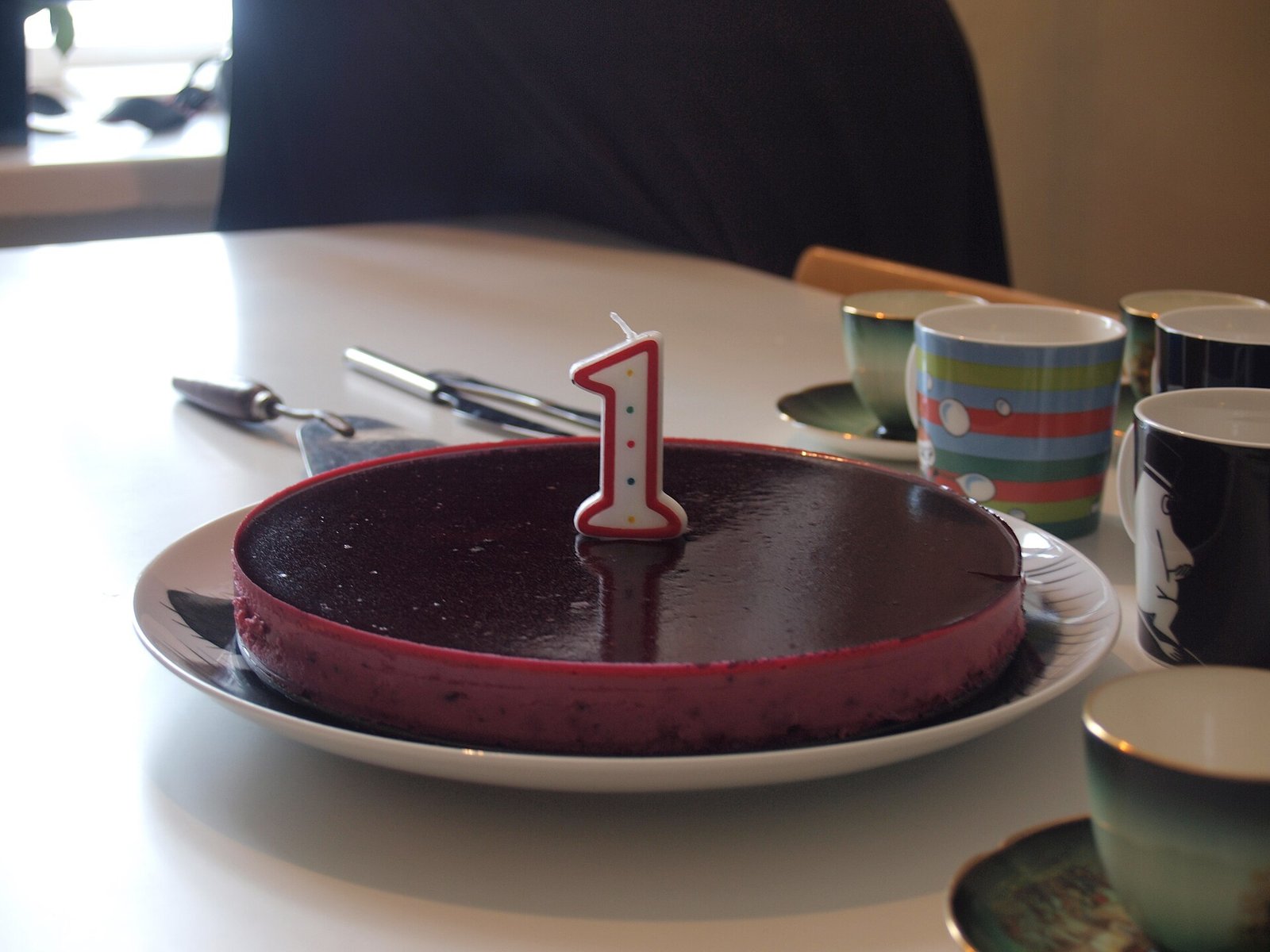 A first-birthday cake with a number-one candle on a table with coffee cups