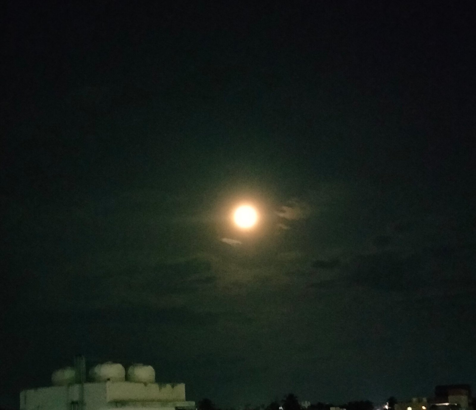 A bright full moon glowing in a hazy night sky above city rooftops.