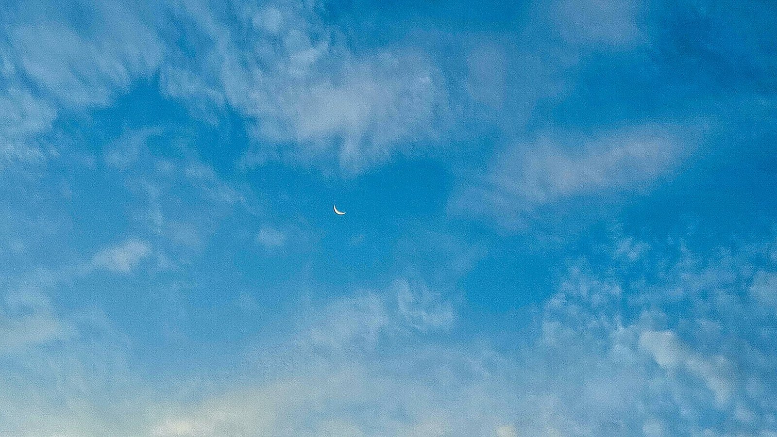A thin crescent moon visible in a blue daytime sky with wispy clouds.