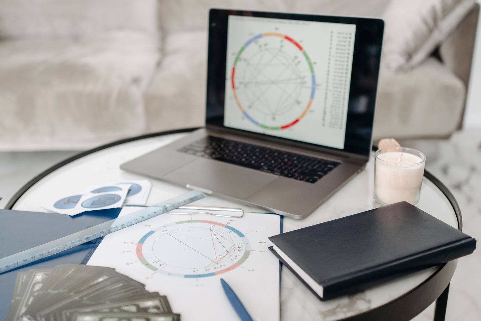 A laptop showing a natal chart wheel on a marble table with a printed chart, ruler, tarot cards, zodiac symbol cards, a notebook, and a lit candle.