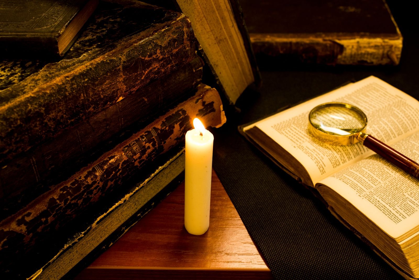 A stack of worn leather-bound books beside a lit candle, with an open book and a magnifying glass.