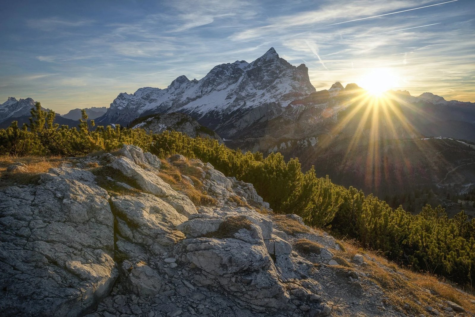 Sun rising over snow-capped mountain peaks above a rocky ridge