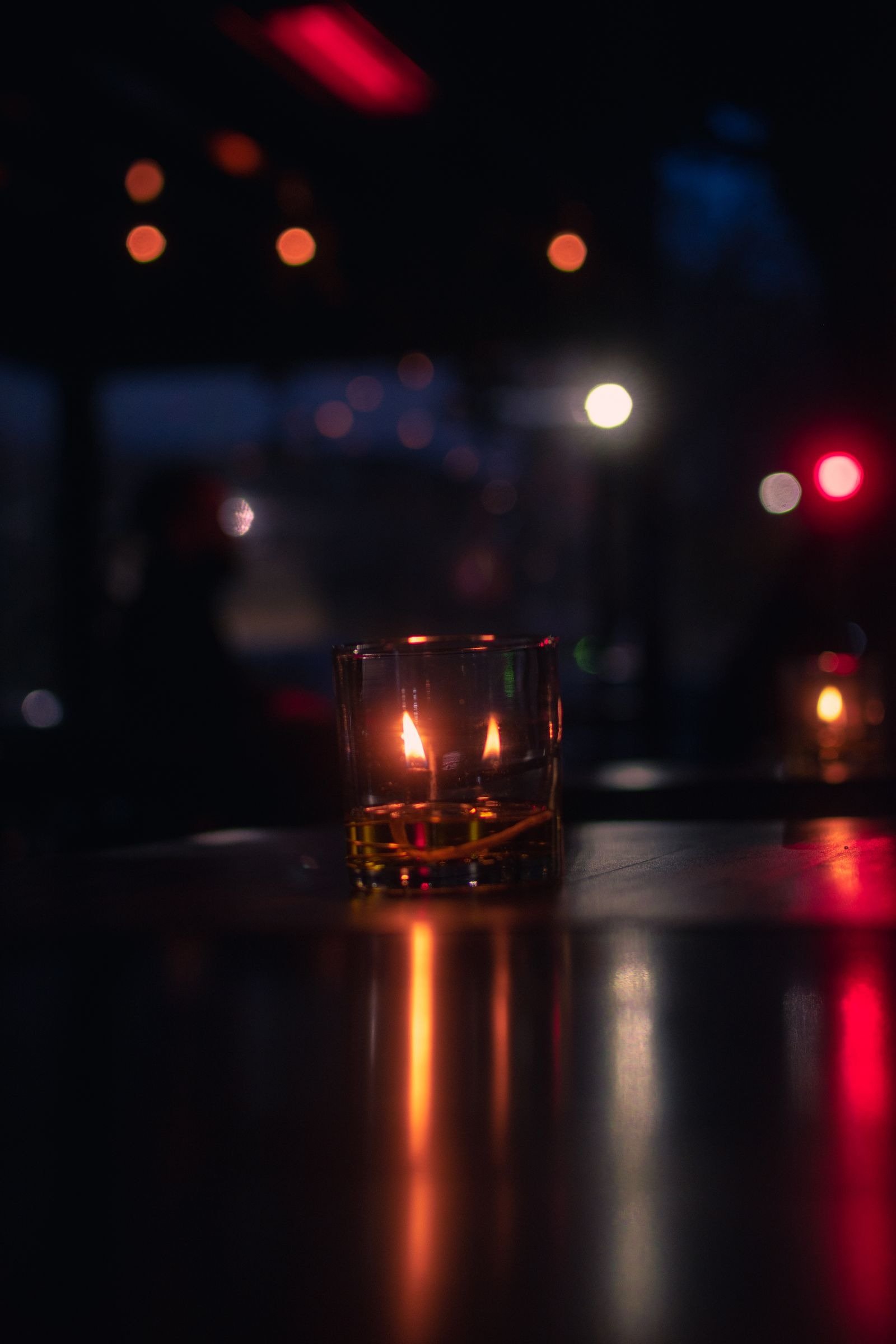 A lit candle in a glass holder on a dark table, with blurred city lights behind.
