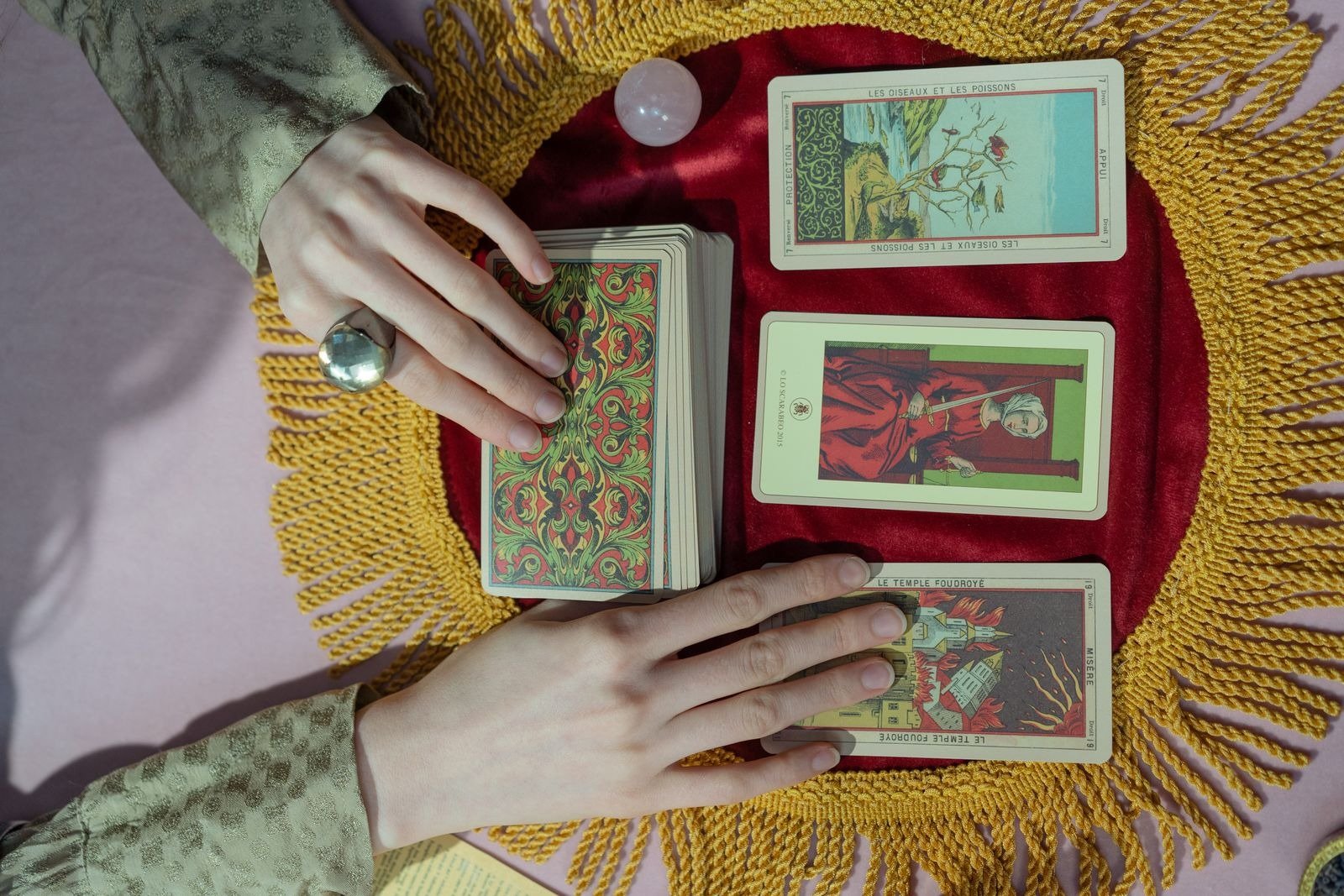 Two hands holding a tarot deck over a spread of cards on a red and gold cloth.