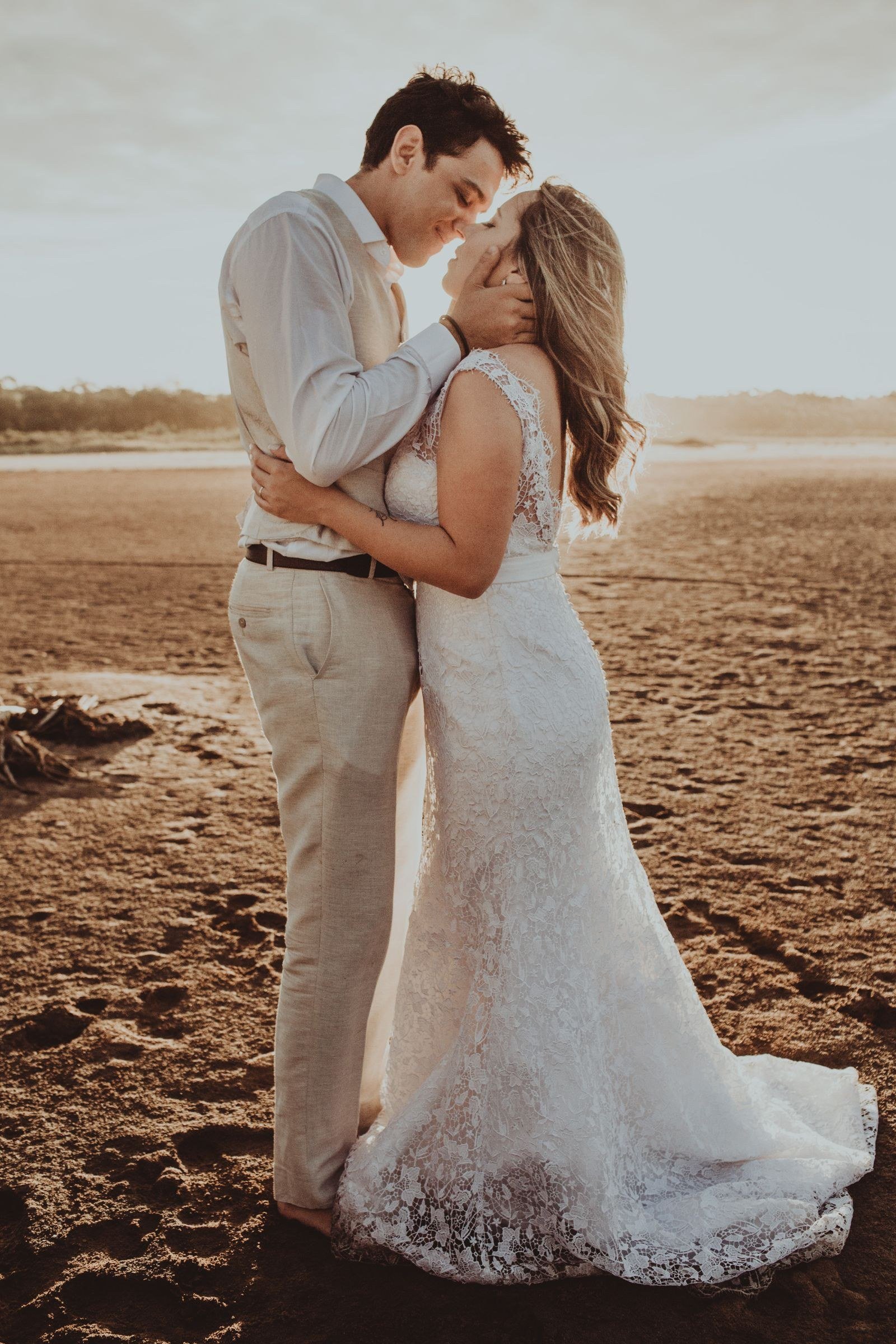 A newly married couple embracing on a beach at sunset, the groom holding the bride's face as they lean in close.