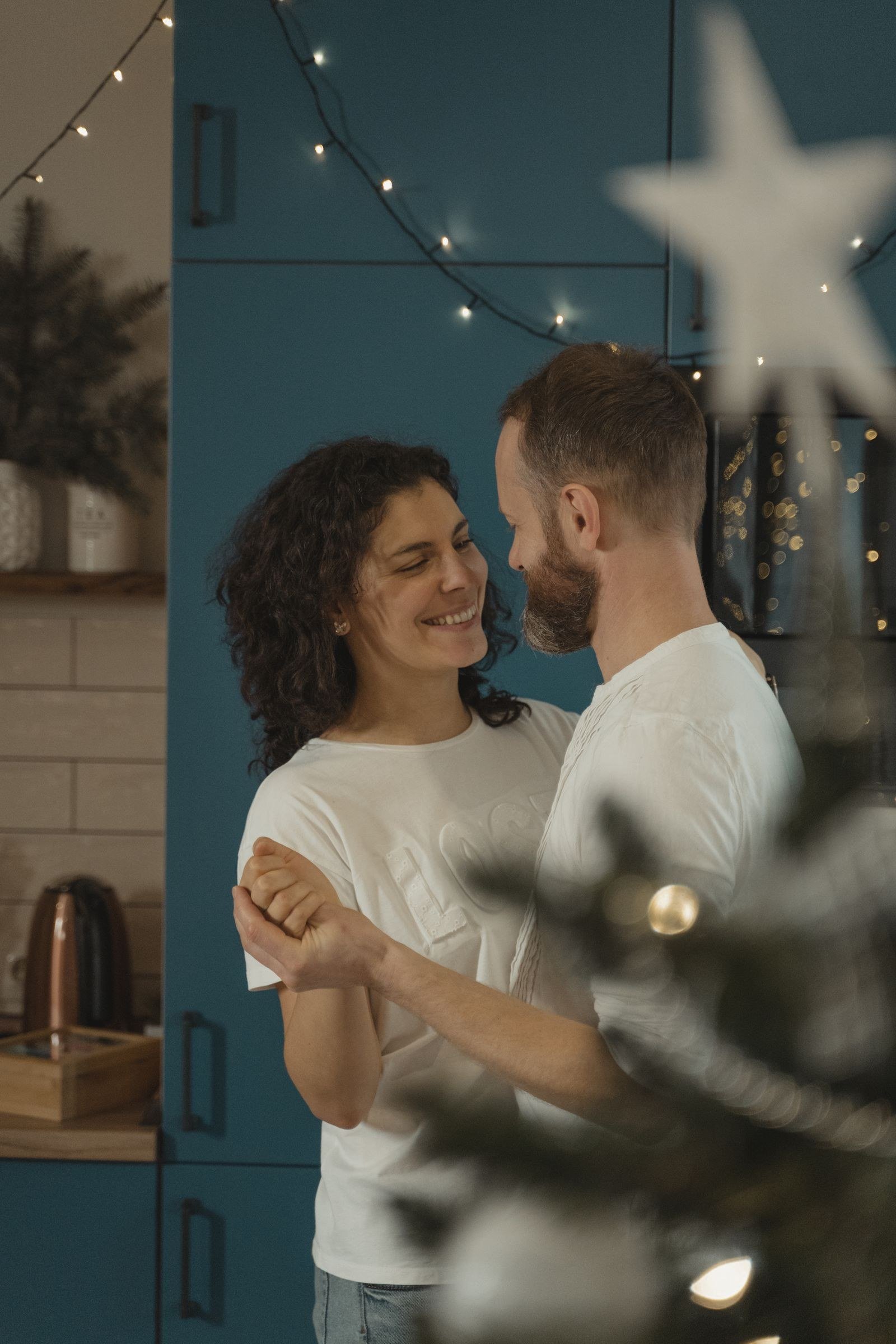 A smiling couple slow dancing in a kitchen decorated with string lights, holding hands.