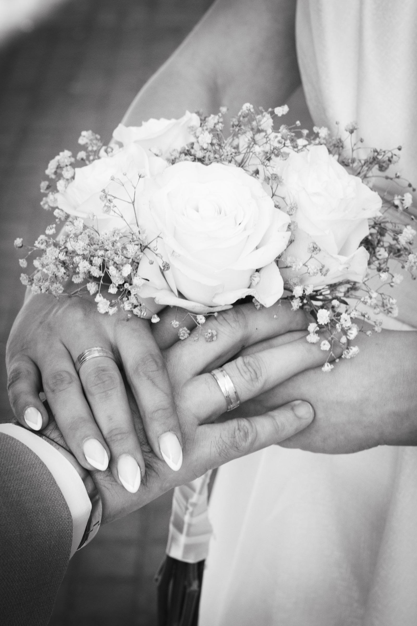 Black-and-white close-up of a couple's hands showing wedding rings, a white rose bouquet resting on them.