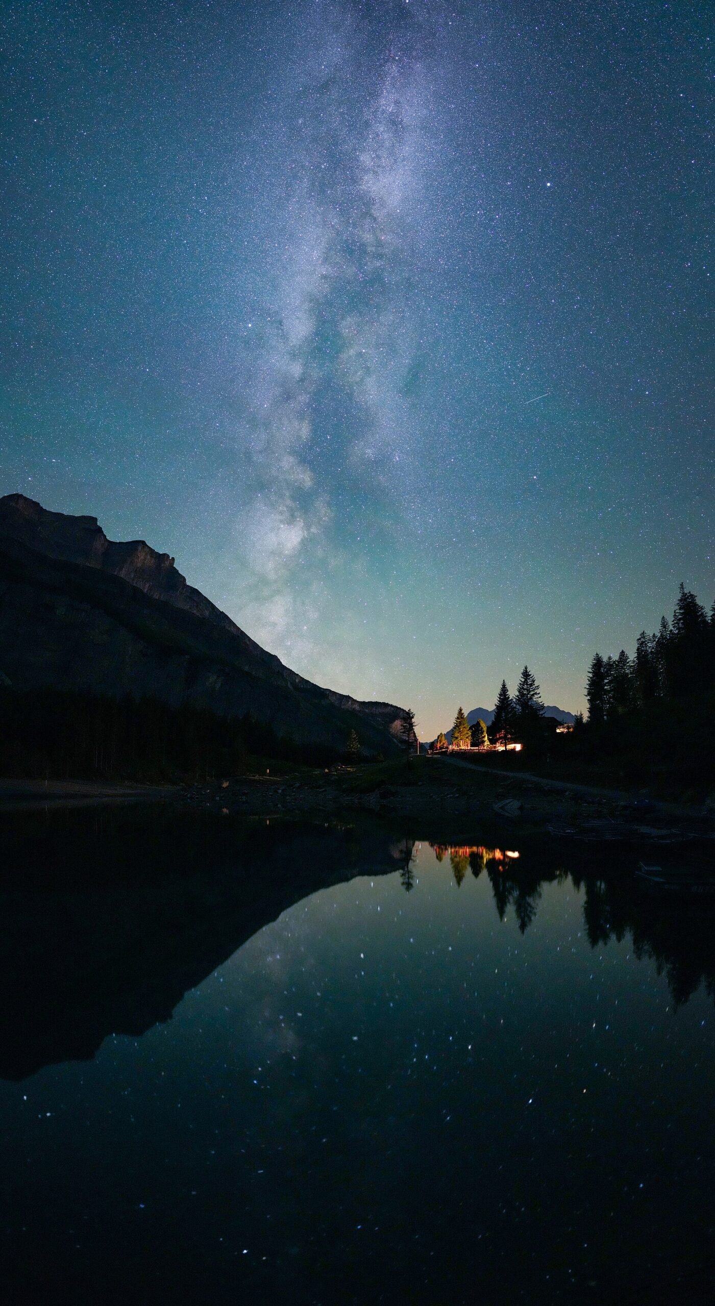 The Milky Way rising over a mountain lake at night, stars and peaks reflected in the still water, a lit cabin on the shore.