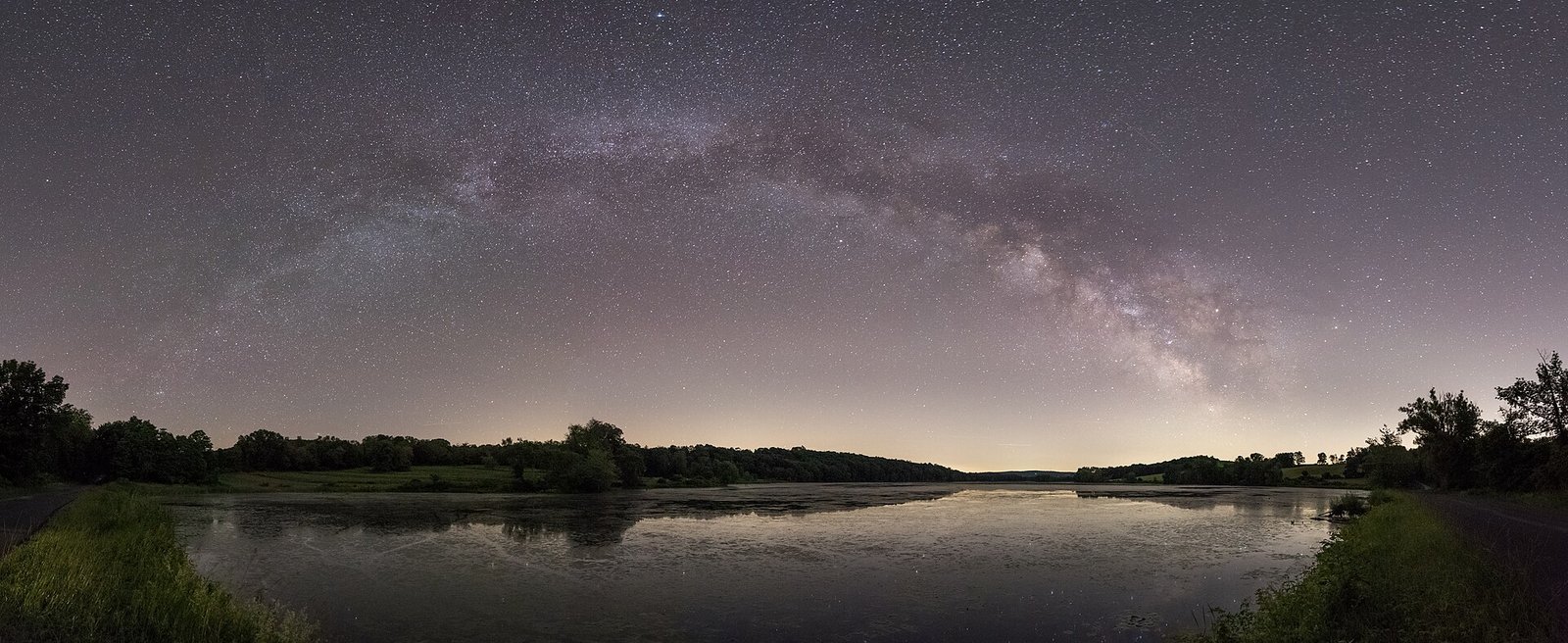 The arch of the Milky Way stretching across the night sky above a calm lake and tree-lined shore.