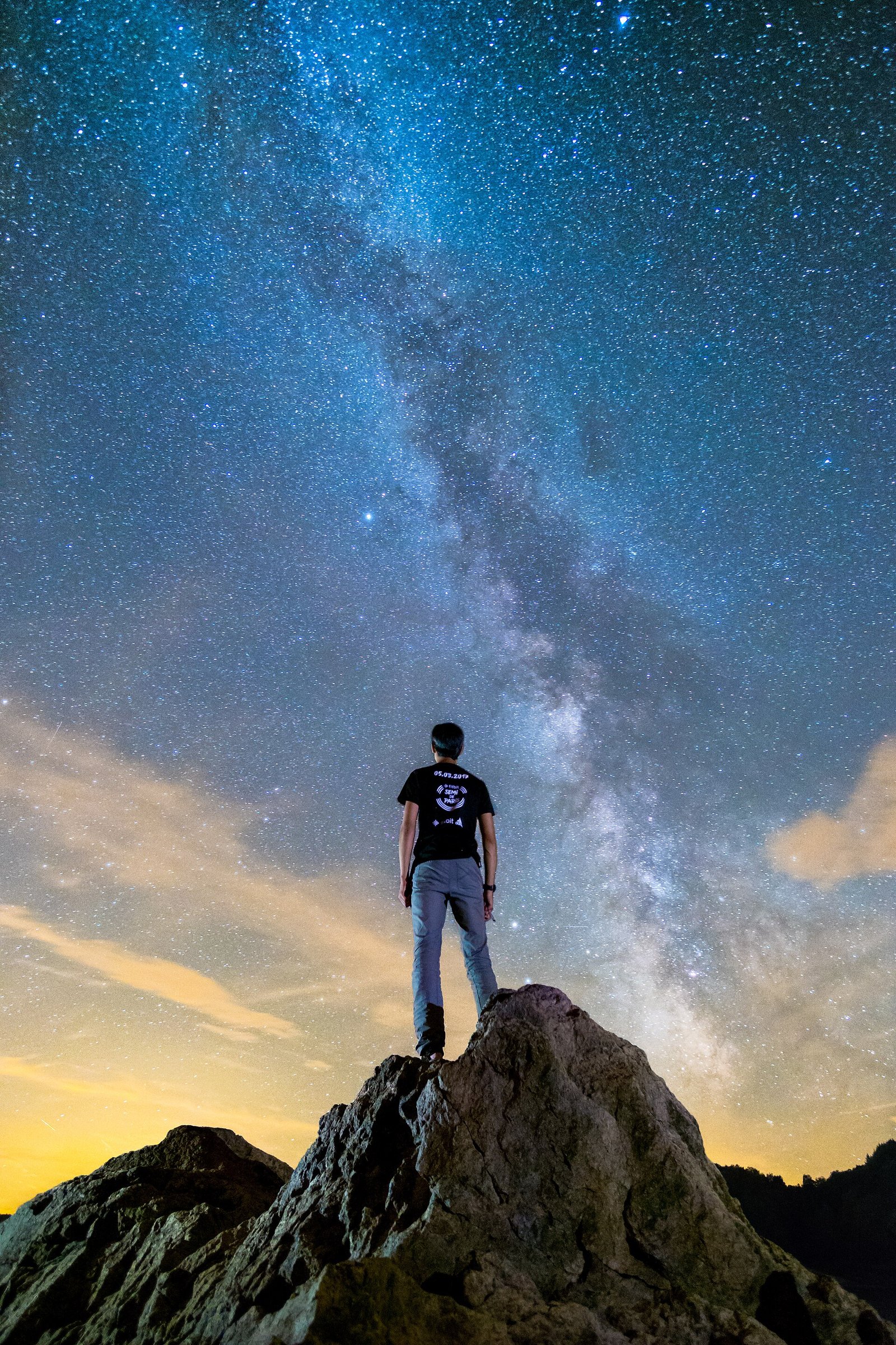 A person standing on a rock at night, looking up at the Milky Way stretching across a deep blue sky.