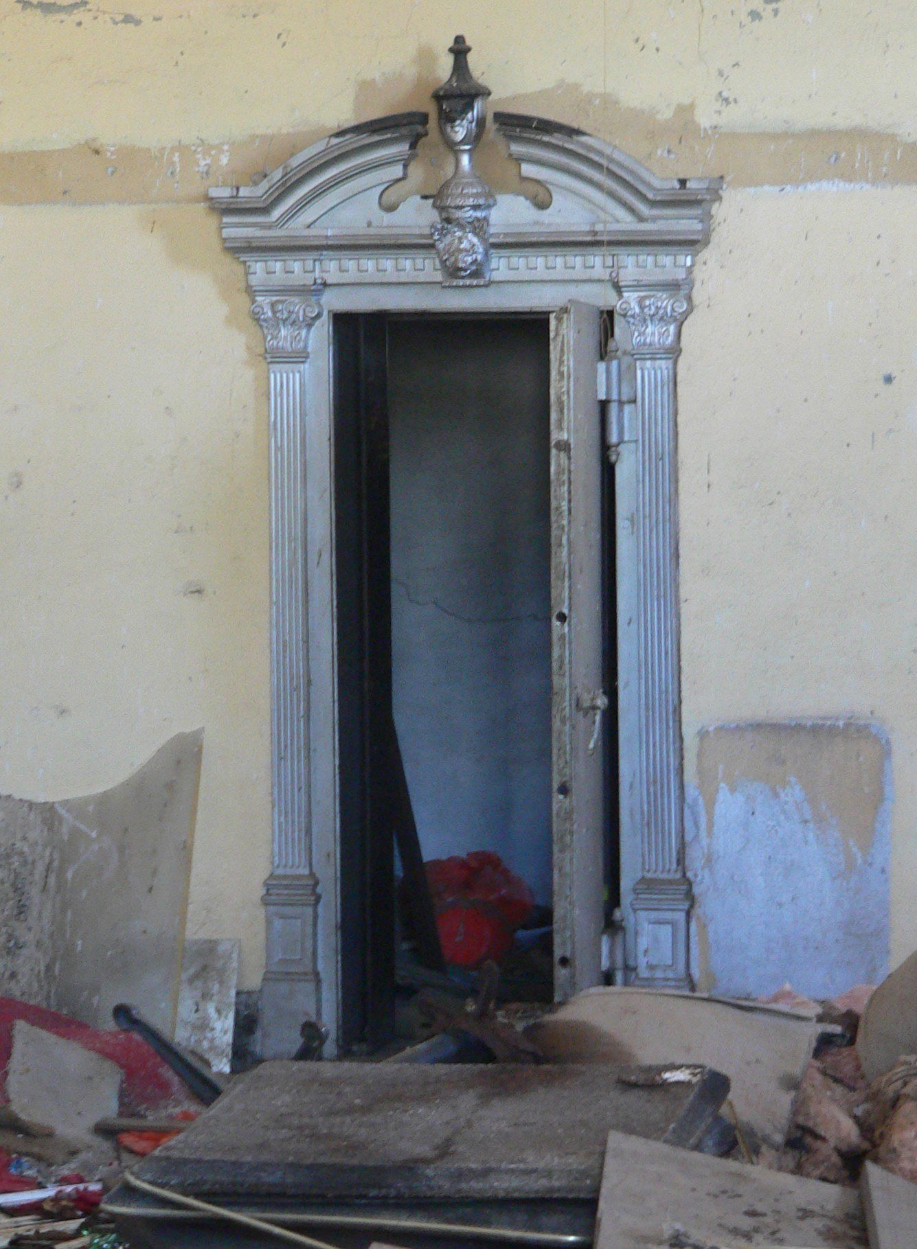 The open steel vault of an abandoned bank in Gage, Oklahoma, framed by fluted columns and a decorative pediment, standing in a derelict room with peeling paint and debris.