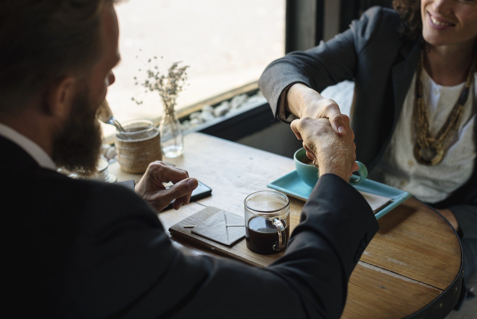 A bearded man and a smiling woman shaking hands across a small cafe table with coffee cups, sealing an agreement.