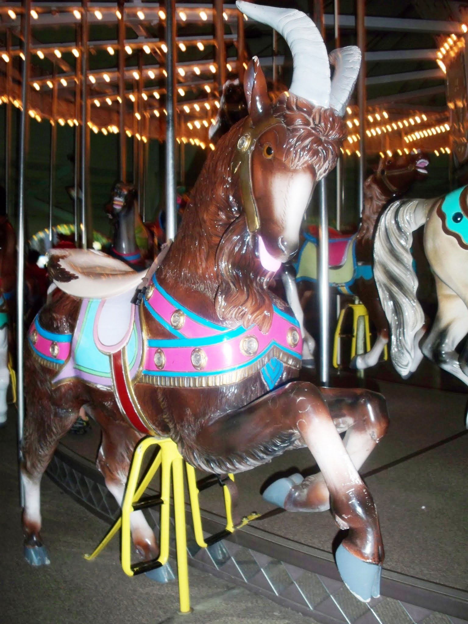 Close-up of a painted carousel horse figure with white horns and a colorful saddle, under strings of lights at night