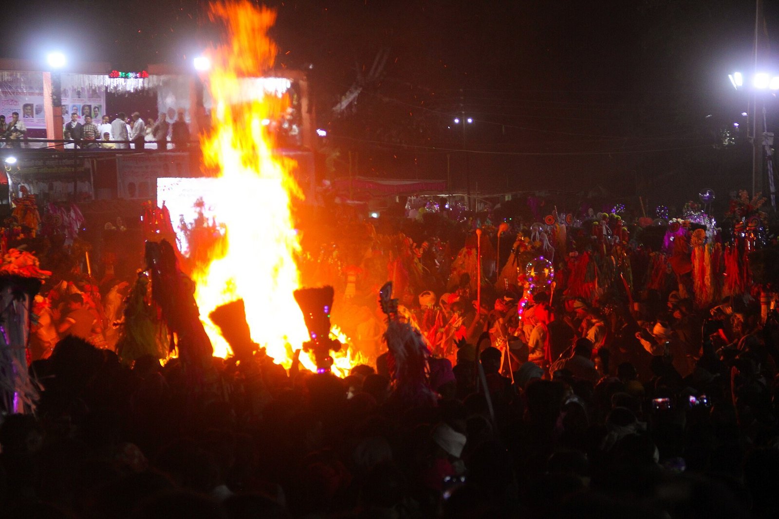 A large bonfire blazing at a nighttime festival, a dense crowd gathered around the flames with colorful decorations and bright lights.