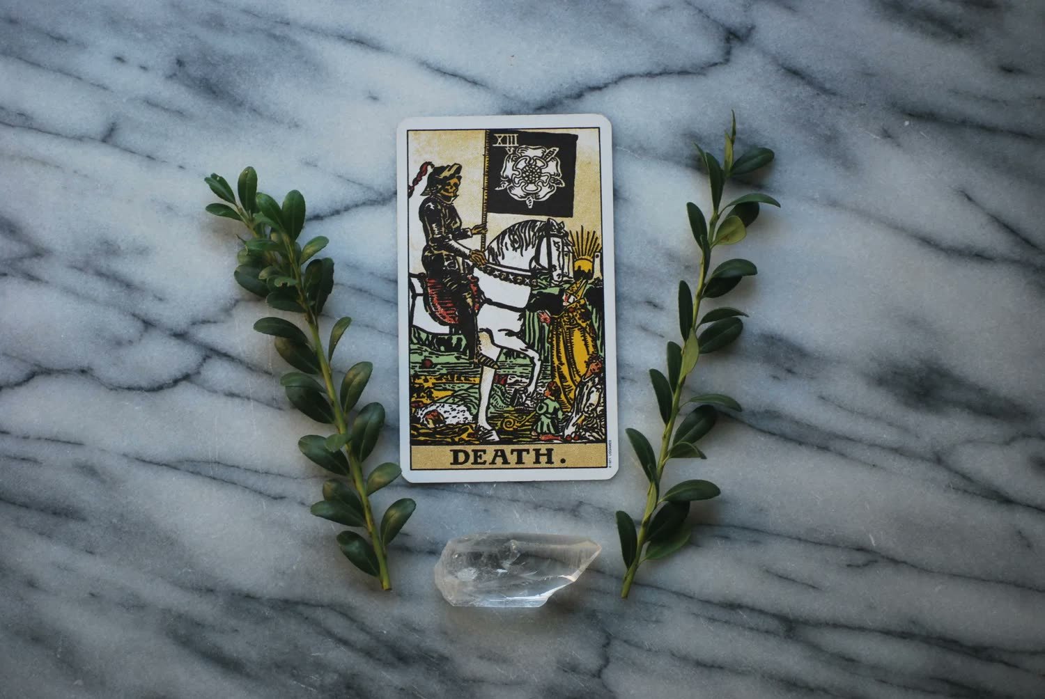 The Death tarot card on a marble surface with green sprigs and a crystal