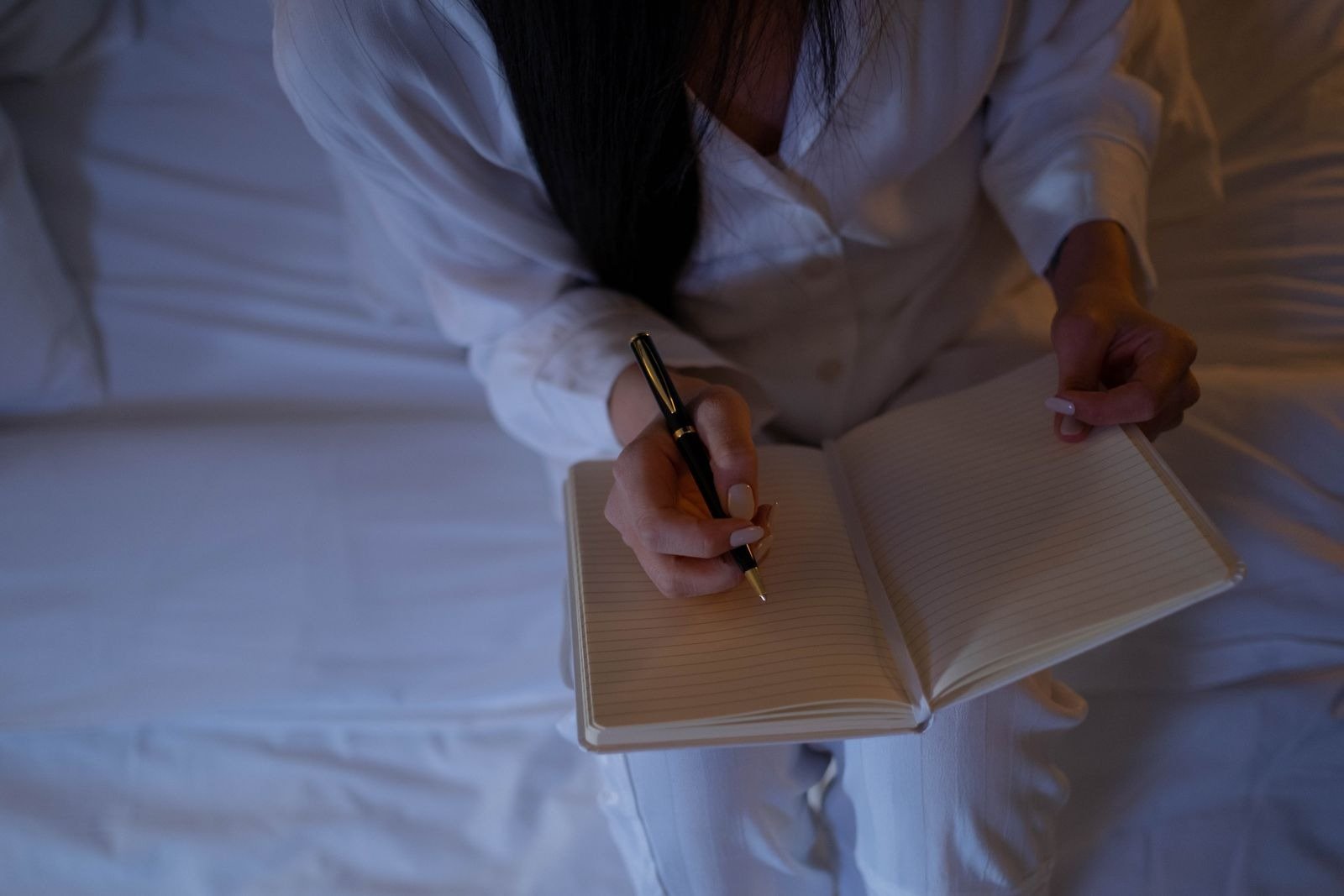 A woman in a white shirt writing in an open journal while sitting on a bed.