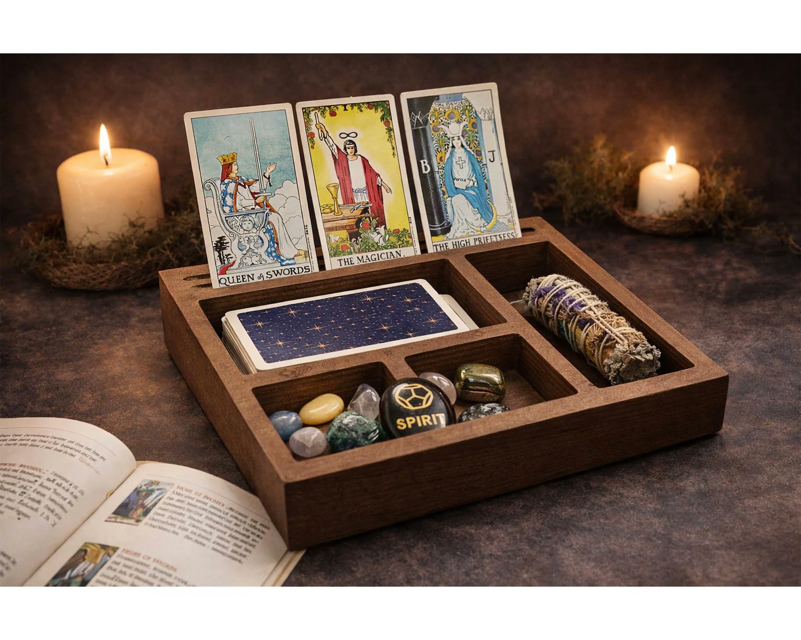 A wooden tarot card holder with three cards displayed, a deck, crystals, and a sage bundle in its compartments, with lit candles behind.