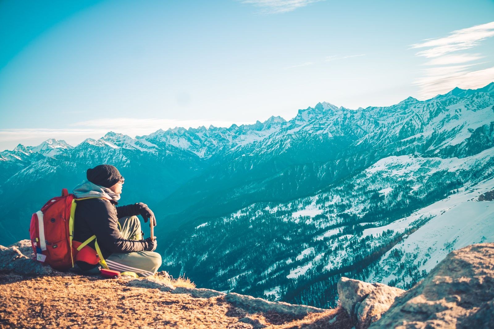 Hiker with a red backpack sitting on a mountain peak overlooking snow-capped ranges.