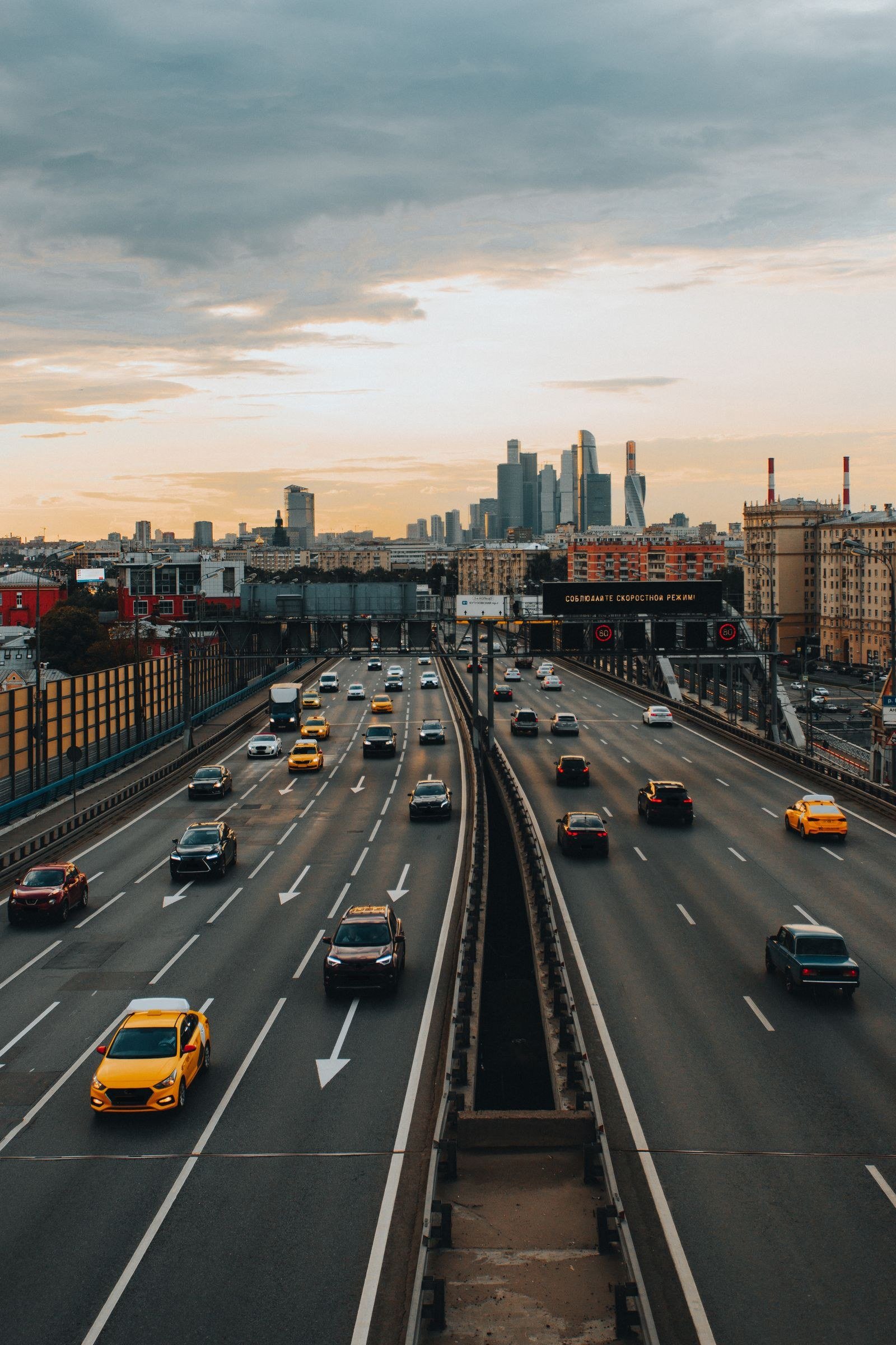 An aerial view of a multi-lane highway at dusk, streams of cars moving through a city under a cloudy evening sky.