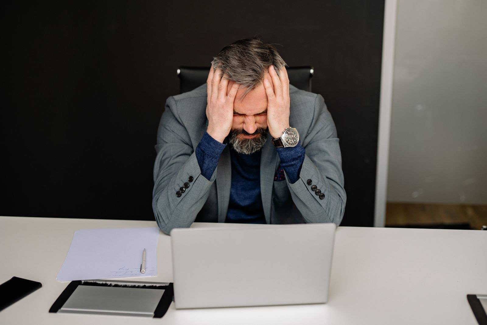 A frustrated young man sitting at a desk with his head in his hands, a laptop open in front of him and papers scattered around.