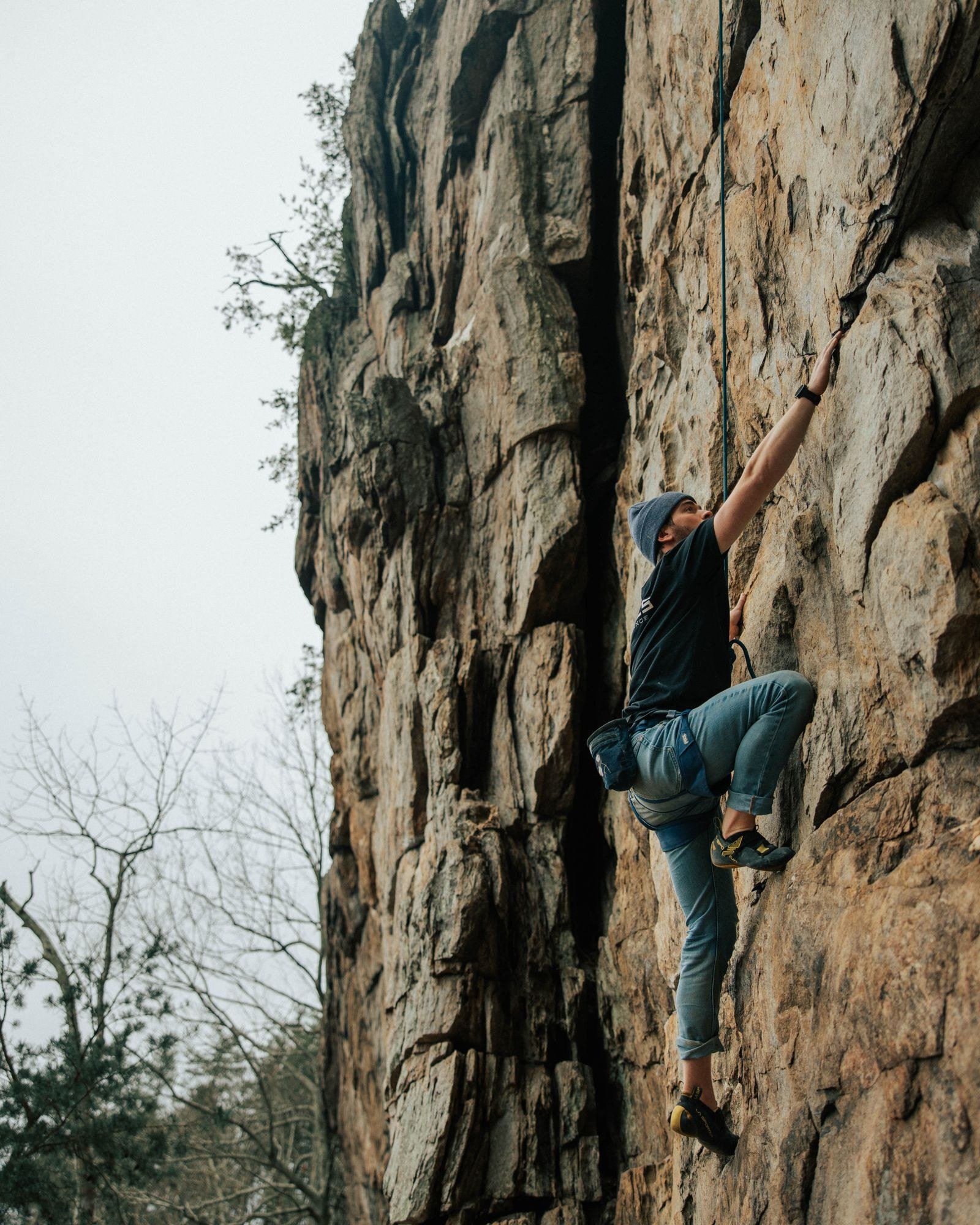 A rock climber in a harness scaling a steep cliff face, chalked hands gripping a narrow hold high above the ground.