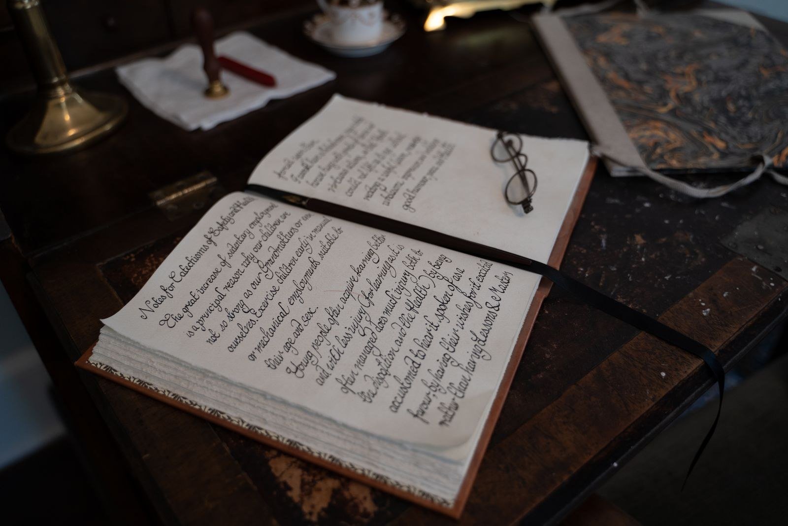 An open book filled with handwritten script on a dark wooden desk, reading glasses resting on the page, a brass lamp and teacup nearby.