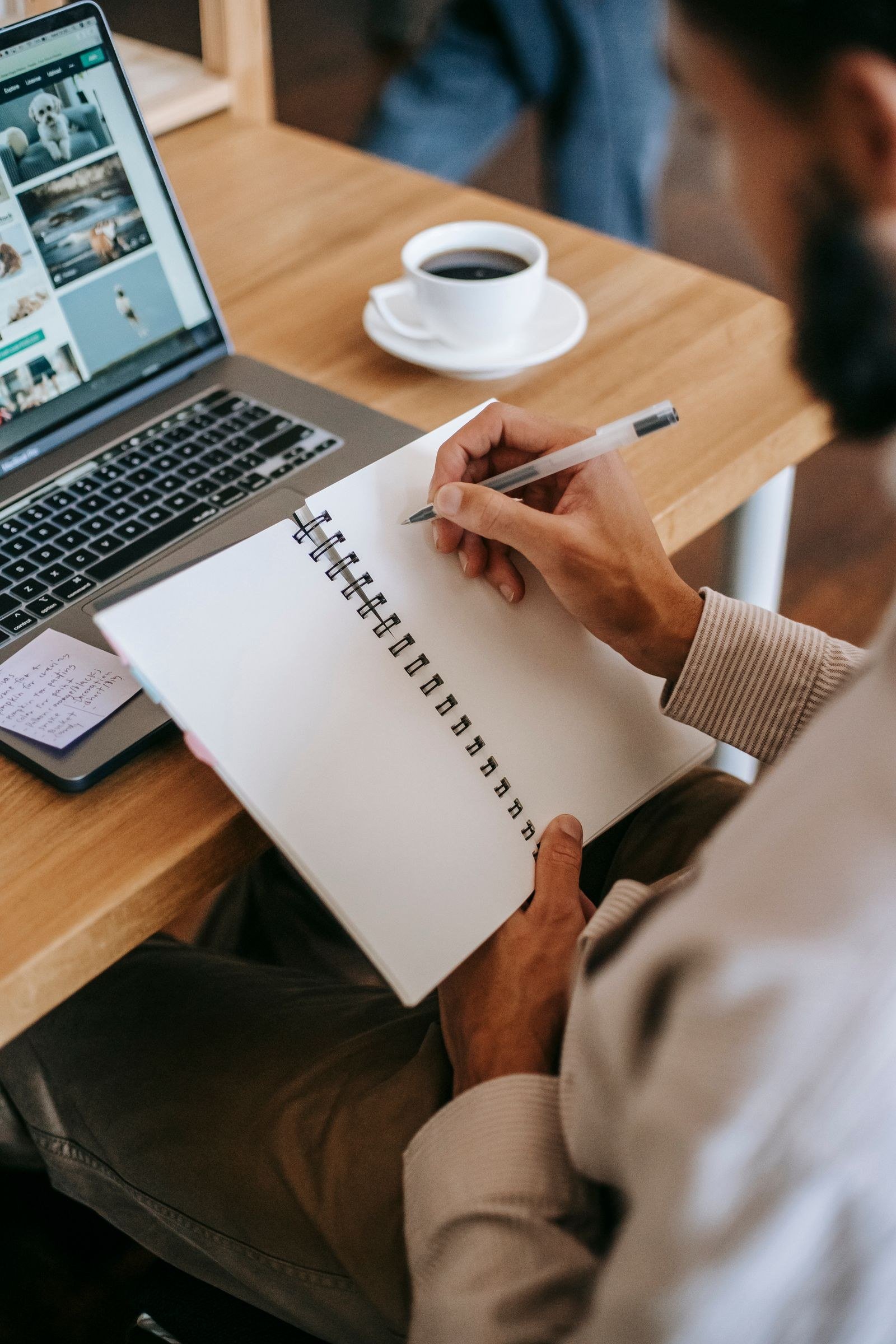 A hand writing in a spiral notebook with a pen, a laptop and a cup of coffee on the wooden table beside it.
