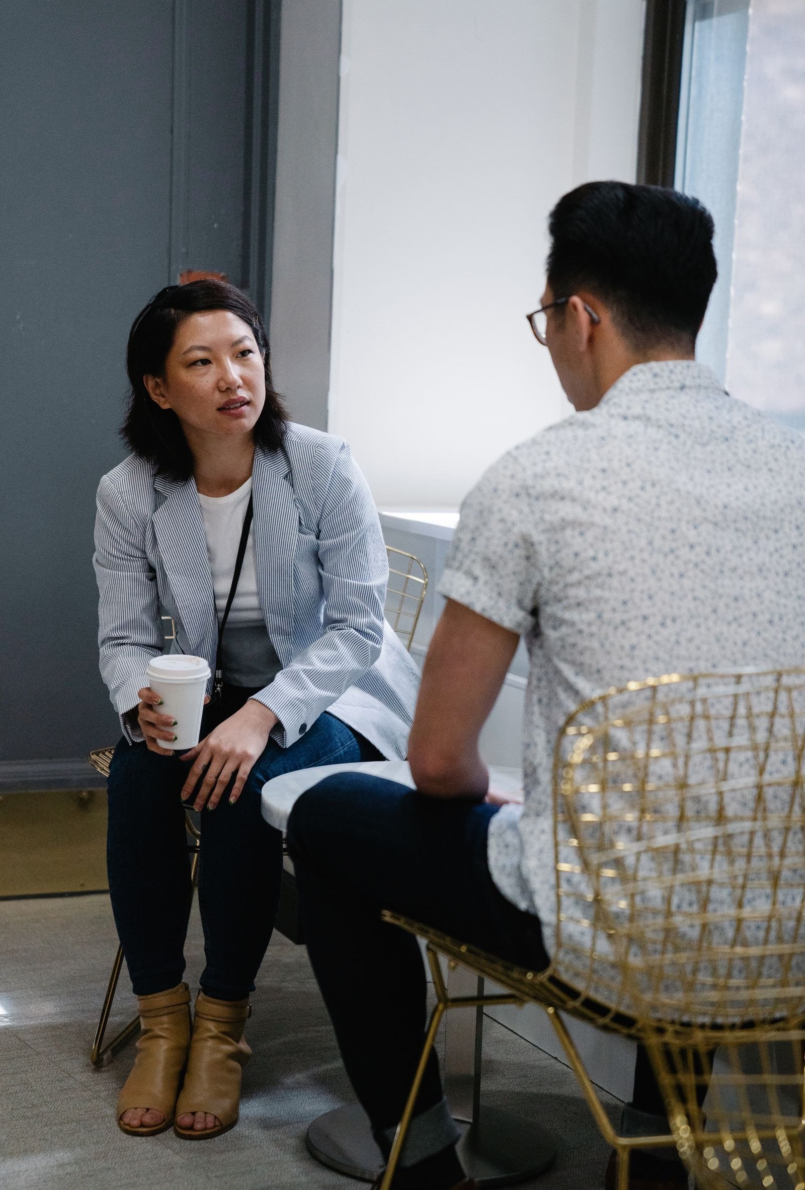 A woman holding a takeaway coffee cup talking with a man at a small indoor table.