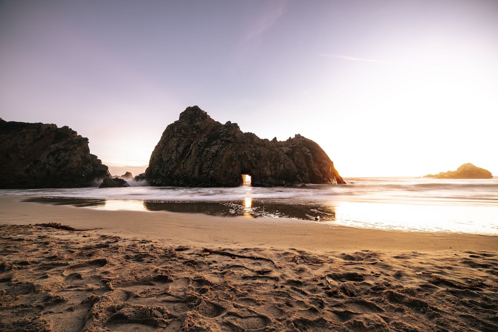 Ocean waves washing around a natural rock arch on a sandy beach at golden hour.