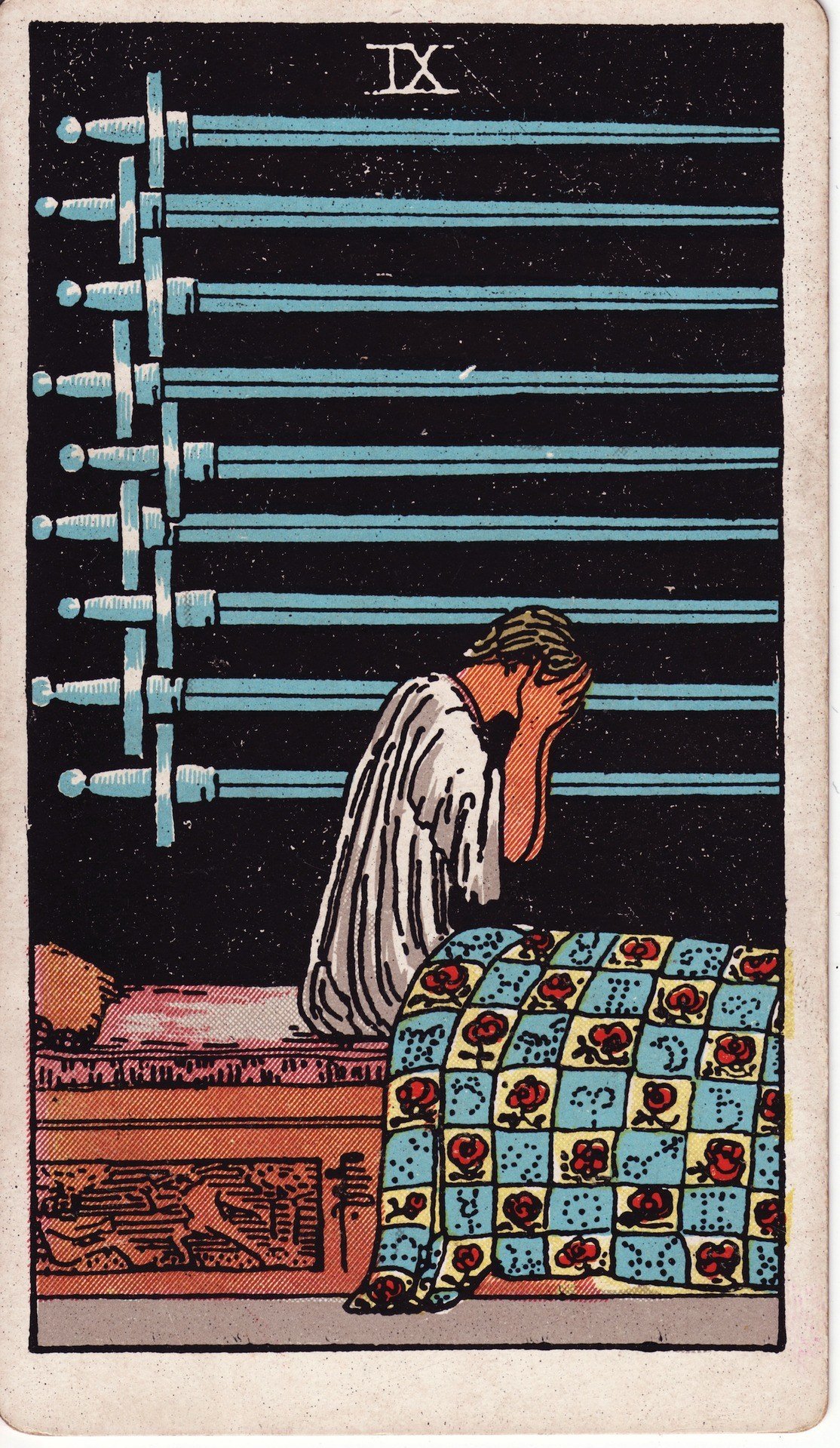 The Nine of Swords from the Rider-Waite-Smith tarot deck: a figure sits up in bed at night with head in hands, nine swords hanging horizontally on the dark wall behind, an quilt patterned with roses and astrological symbols.