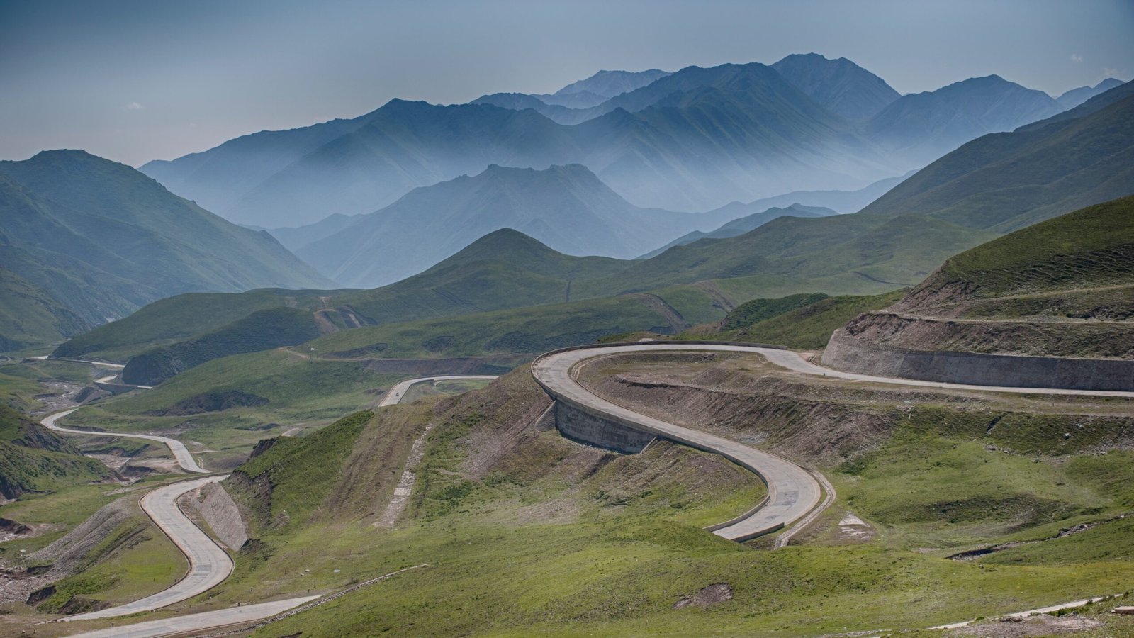 Winding paved road switchbacking through green mountain hills under a hazy sky