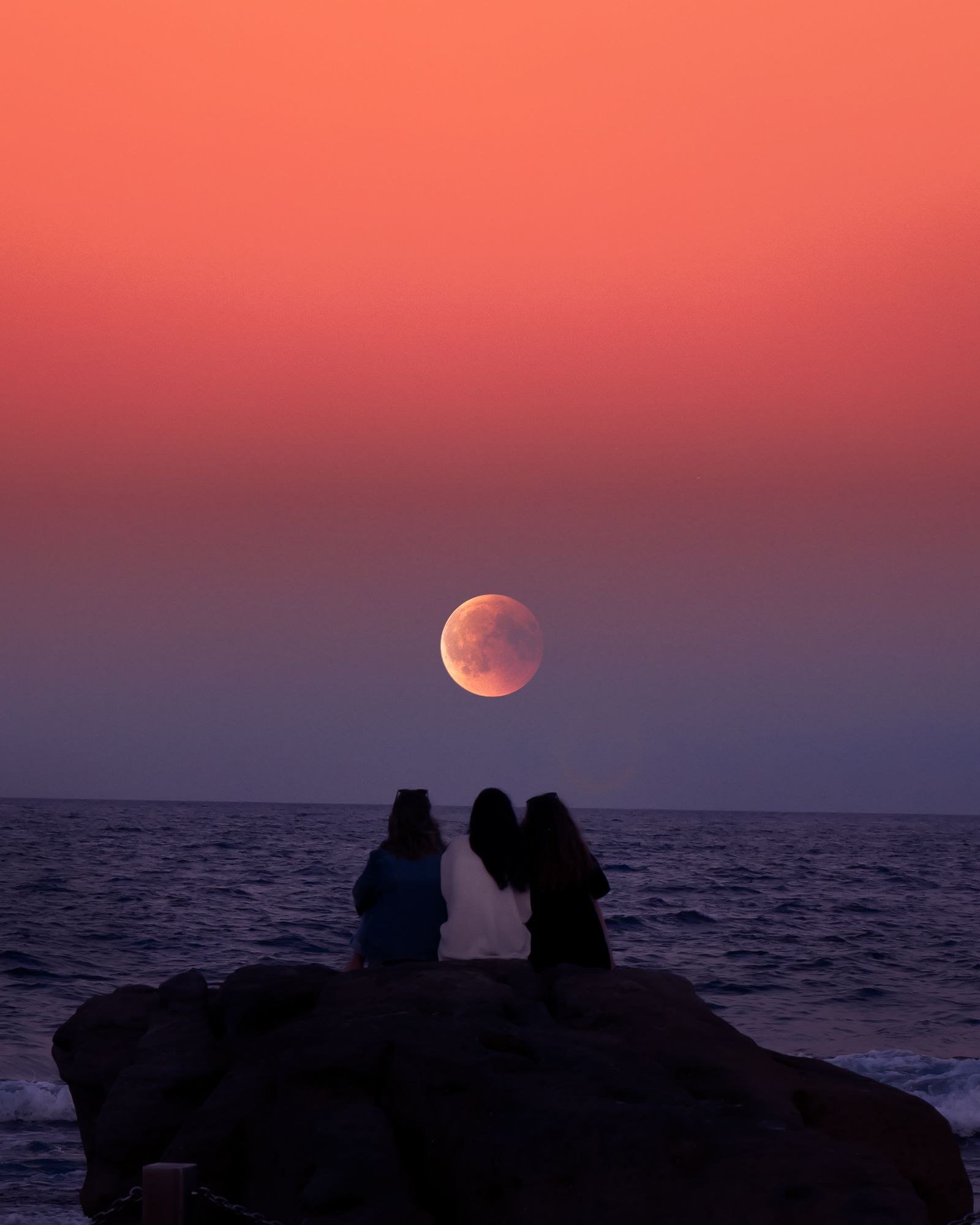 Three people sitting on a rock by the ocean watching a large full moon rise at dusk.