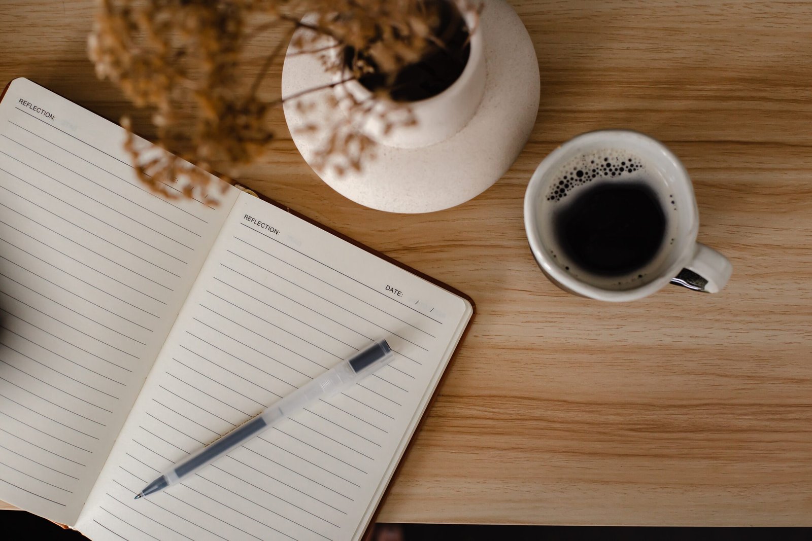Overhead view of an open journal with a pen, a cup of coffee, and a vase of dried flowers on a wooden table