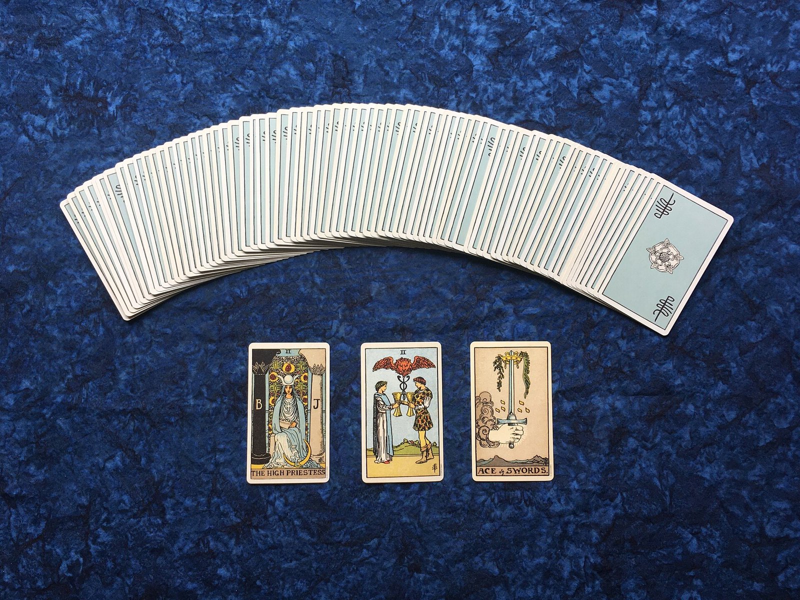 A fanned Rider-Waite tarot deck with three cards face up (High Priestess, Two of Cups, Ace of Swords) on blue cloth