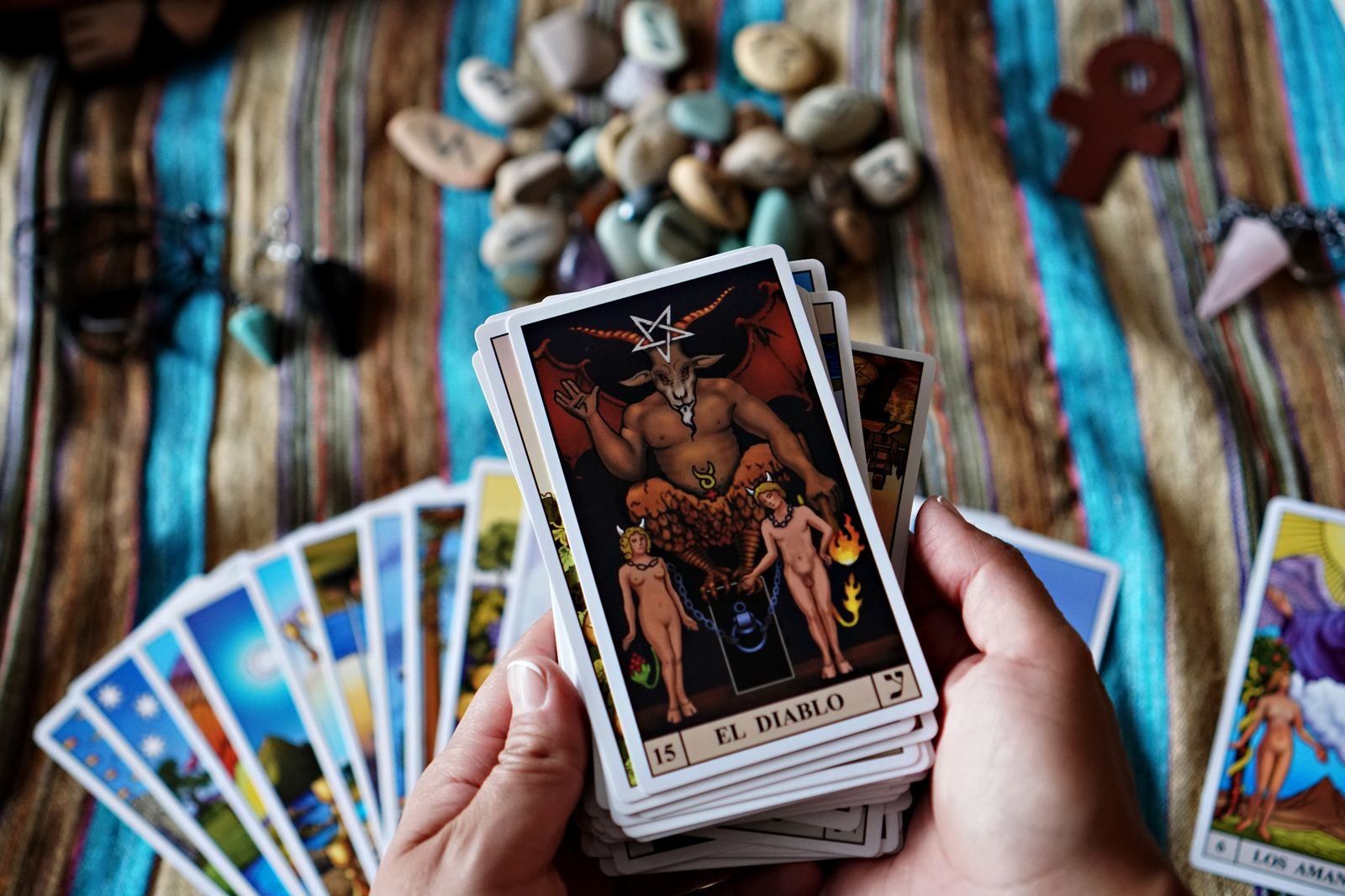 Hands holding a tarot deck with the Devil card (El Diablo) on top, over a striped cloth with runes