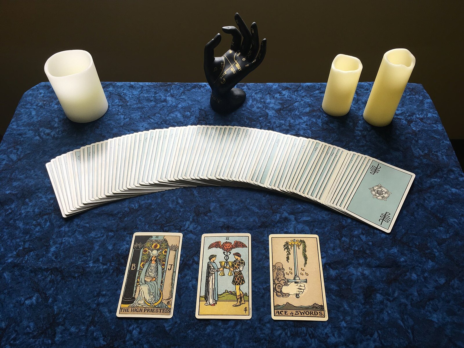 A fanned tarot deck with three face-up cards on blue cloth, candles and a hand-shaped candle holder behind it