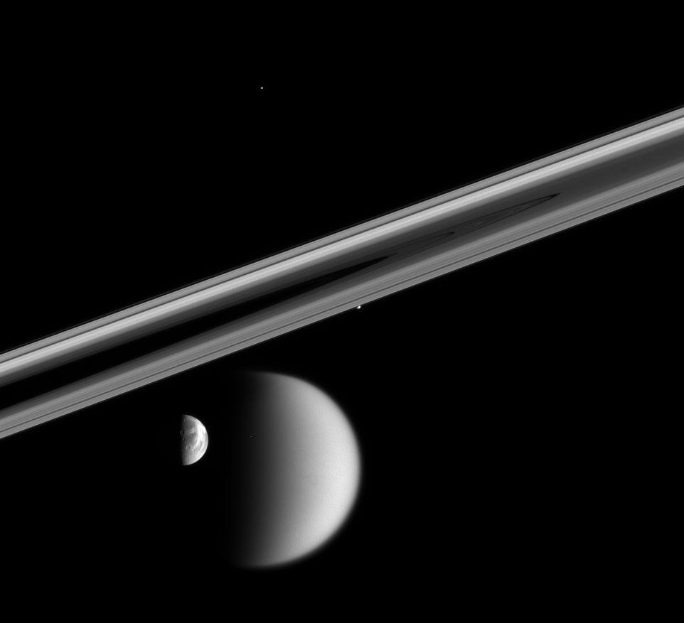 A black-and-white Cassini photograph of one of Saturn's moons passing in front of the planet, with the rings crossing the frame diagonally.