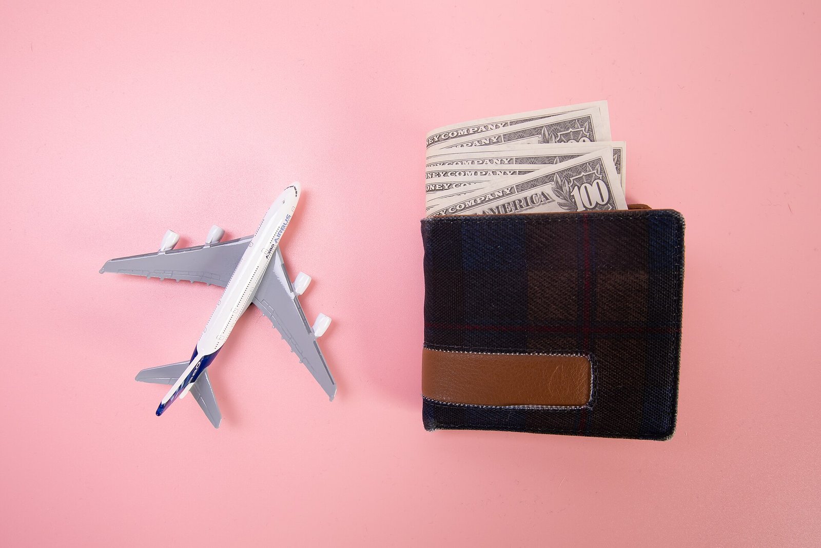 A wallet with cash and a toy airplane on a pink background