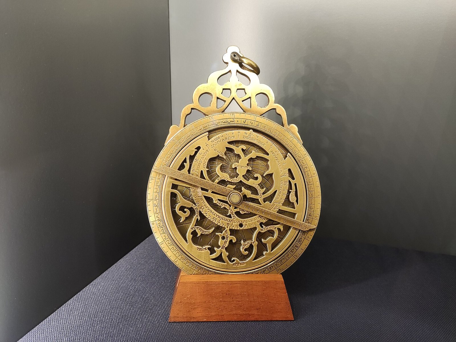A brass astrolabe with an ornate pierced suspension loop, standing on a wooden base in a museum display.