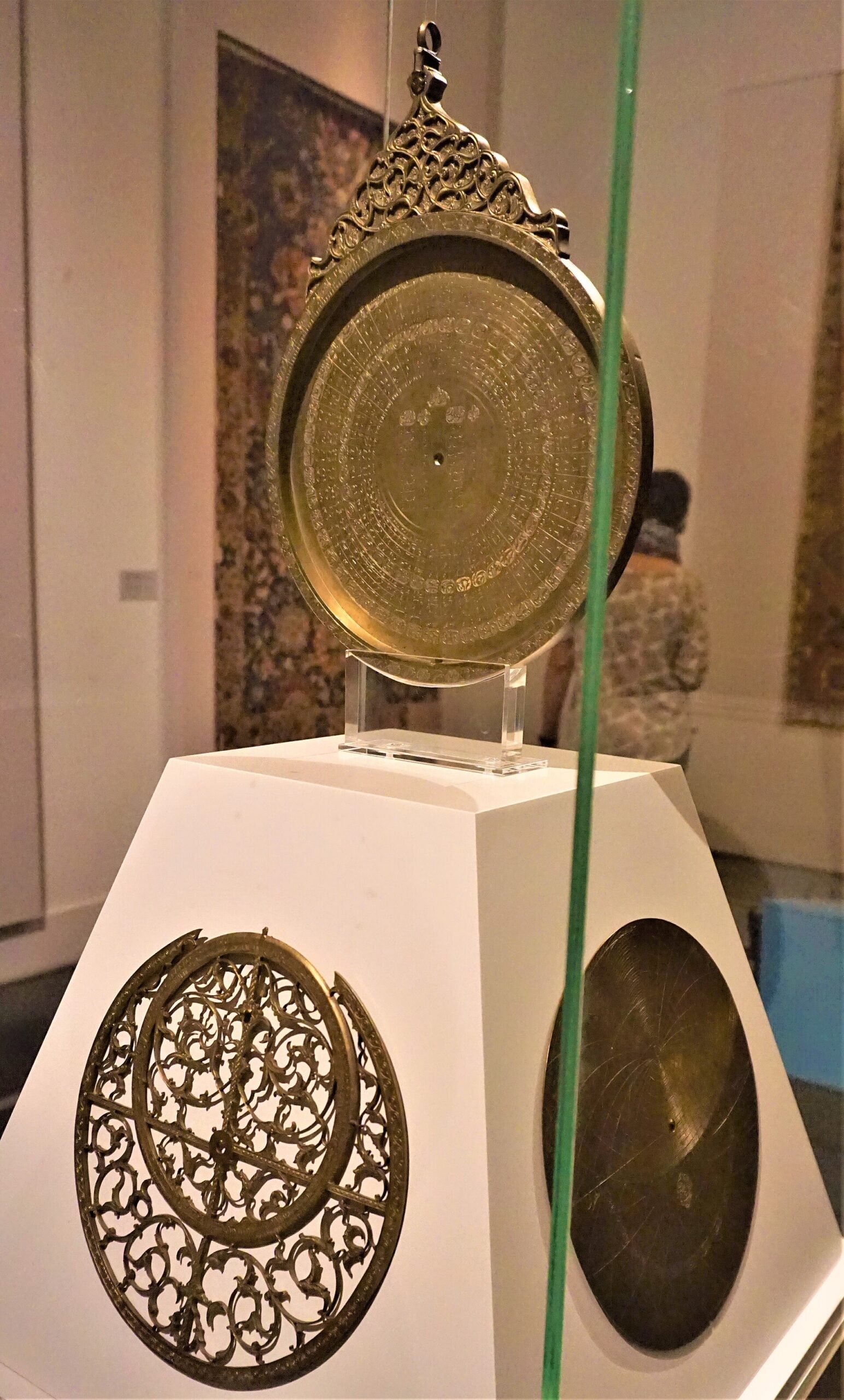 The engraved back plate of an Islamic astrolabe on a museum pedestal, seen through display glass.