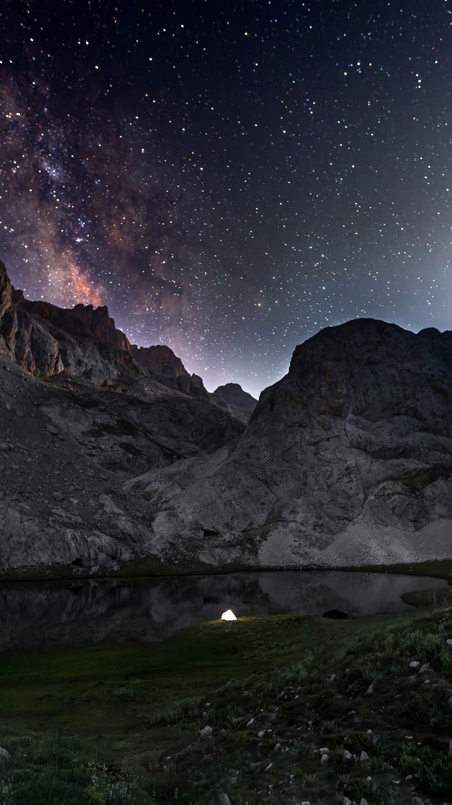 A starry night sky with the Milky Way over rocky mountains, a lake, and a lit tent.