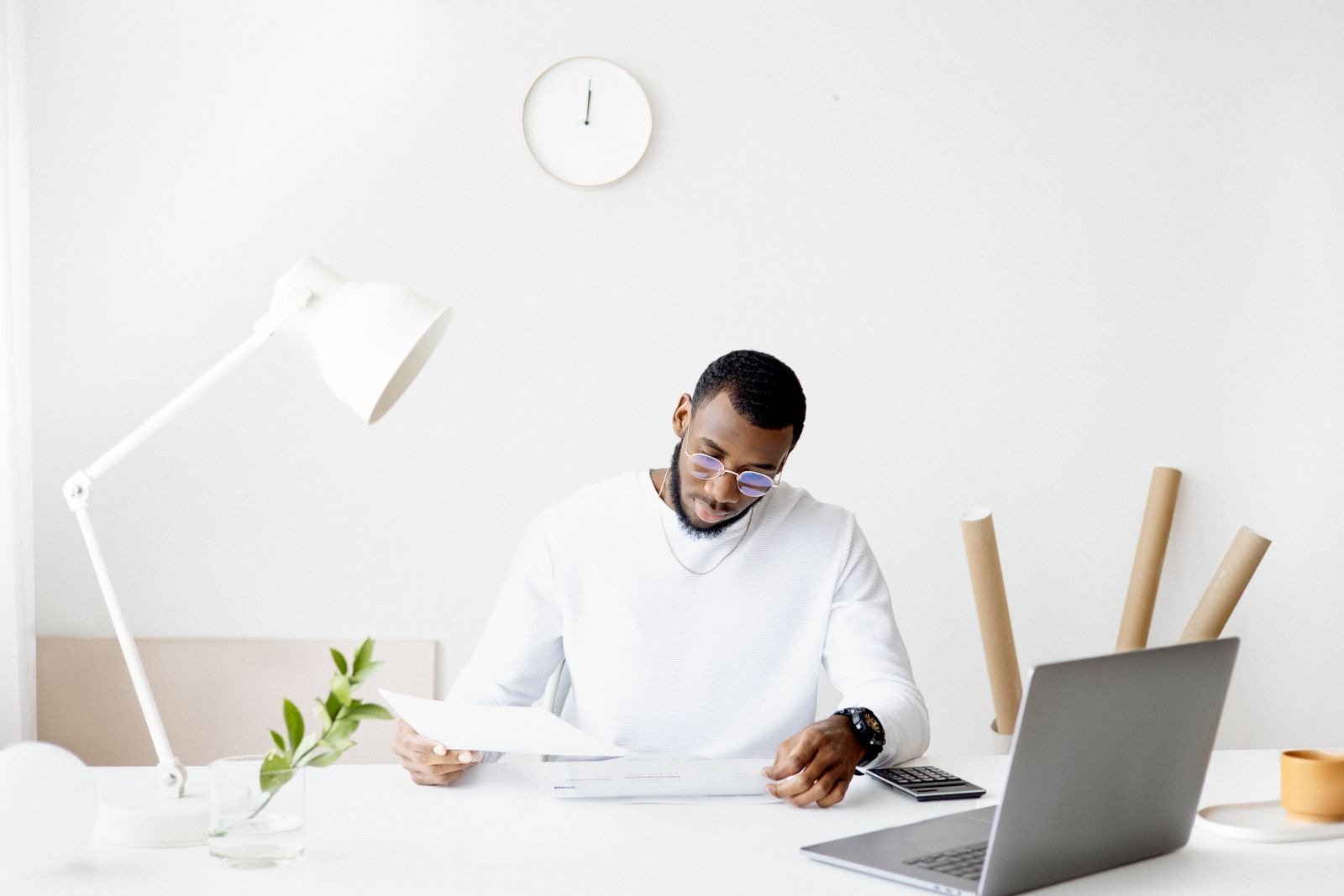 A man reviewing papers at a minimalist white desk with a laptop and desk lamp.